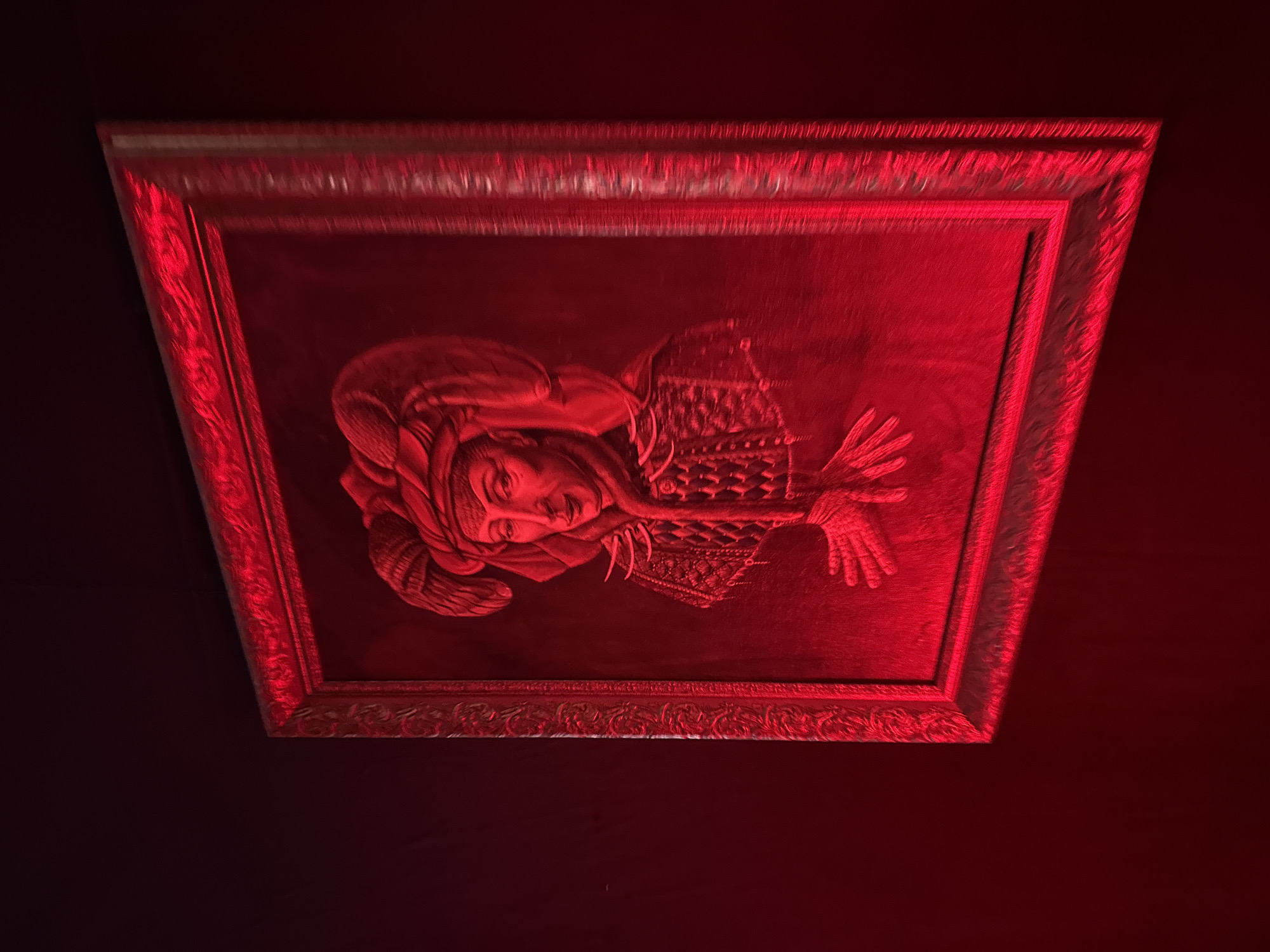 A framed portrait of a woman wearing a head covering and an ornate garment hangs on a dark red wall. The lighting is predominantly red, casting an intense hue over the entire scene. The portrait features a woman with her hands outstretched. The frame is decorative, with raised detailing. The location is Yerevan, Armenia.