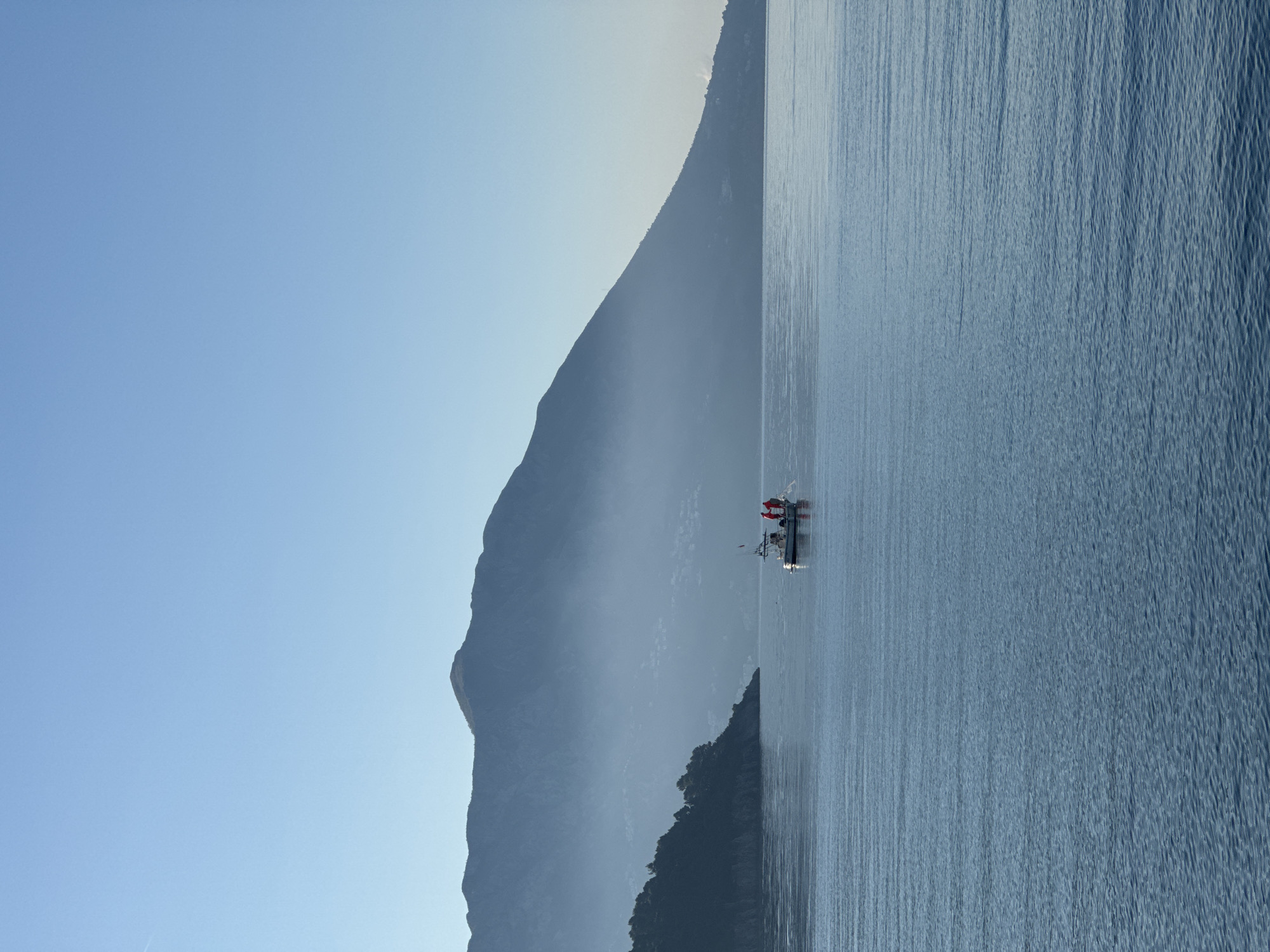 The image shows a lake or sea with a mountain range in the background. A small boat with two people is visible in the water. The sky is clear and blue. The location is most likely in the city of Budva, Montenegro, as the picture contains a shoreline and boats.