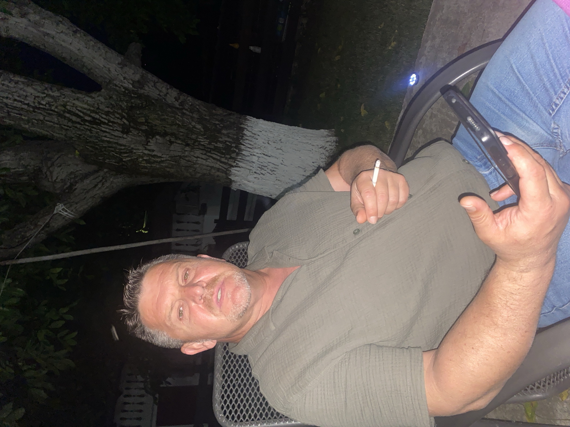 A middle-aged Caucasian man is sitting in a chair outdoors at night. He has short, graying hair, a mustache, and a goatee. He is wearing a green button-down shirt and blue jeans. He is holding a cigarette in his right hand and a smartphone in his left hand. The man is looking at the camera. In the background, there is a tree with white paint on its trunk, and some fencing and foliage. The scene appears to be in a residential area. This location is in Târgu Neamț, Romania.