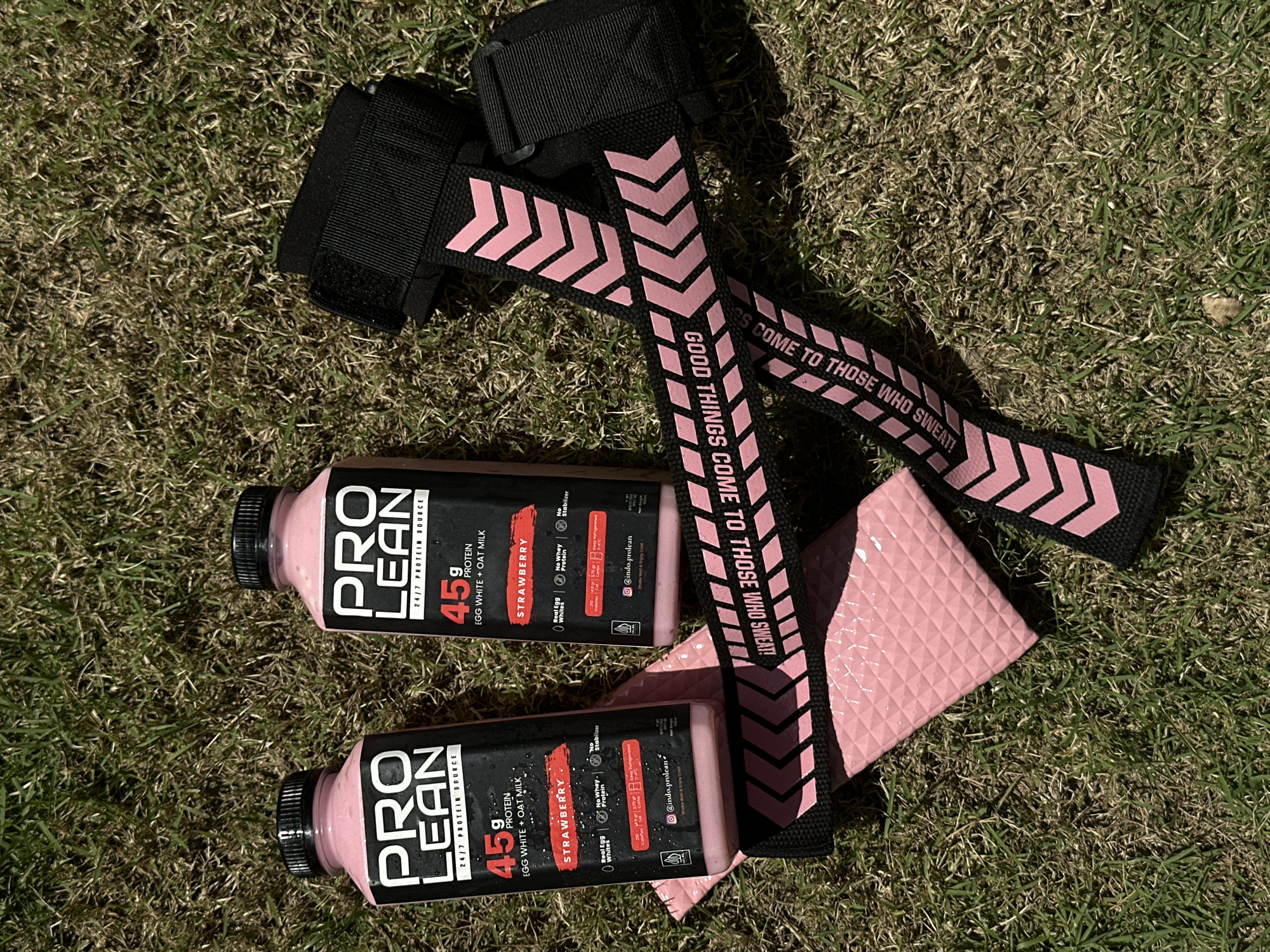 Two bottles of ProLean strawberry-flavored protein drink and a pair of pink and black lifting straps are placed on a patch of dry grass. The bottles are black with white and red lettering. The lifting straps are black with pink chevron patterns and the words "GOOD THINGS COME TO THOSE WHO SWEAT!" printed on them. A pink textured mat is partially visible beneath the straps and bottles. The scene is outdoors, likely on a sunny day given the shadows. This image is not located in North Kuta, Indonesia.