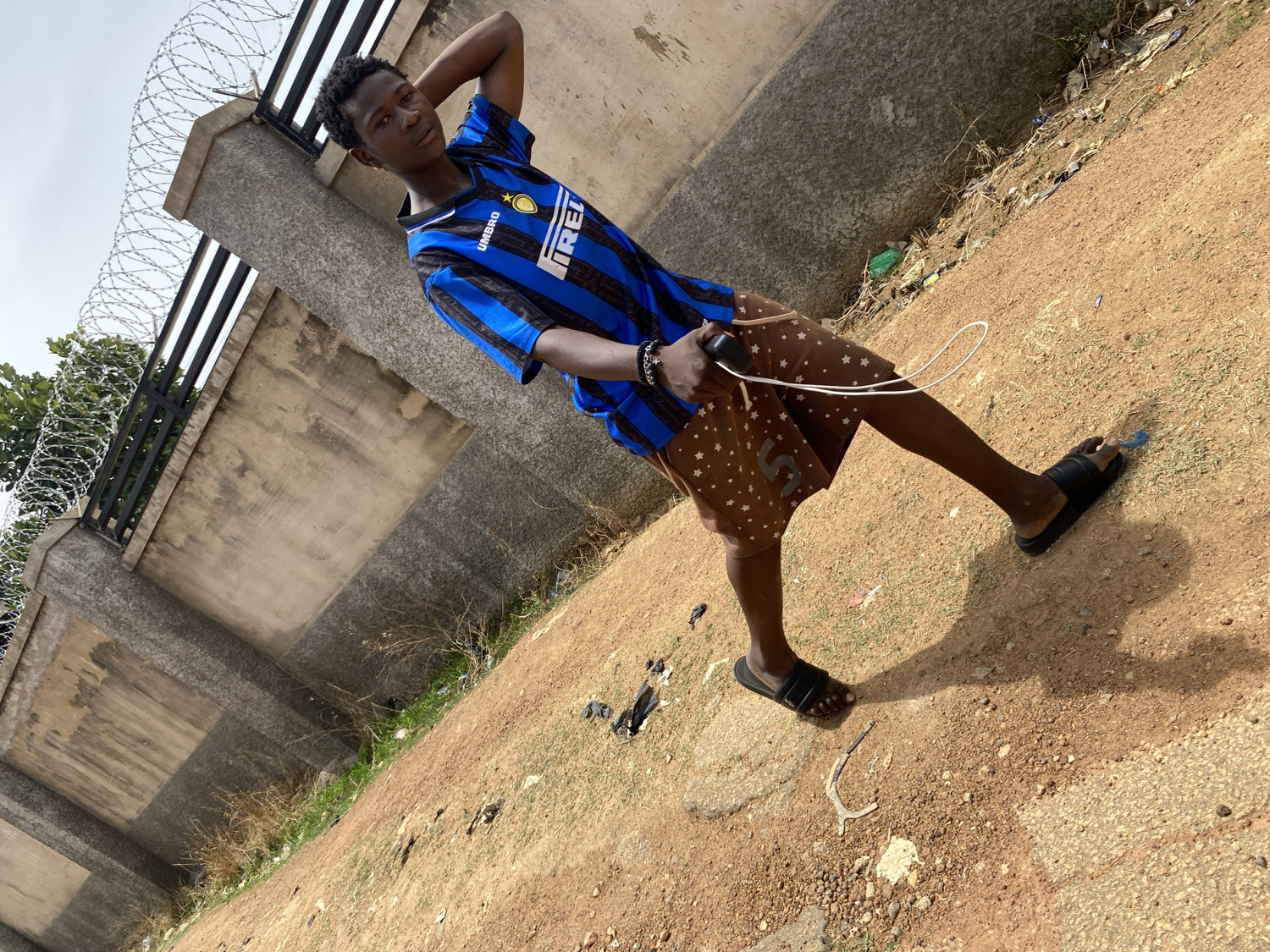 A young man poses for a photograph outdoors in Barakin Akawo, Nigeria. He stands with his left arm bent and hand behind his head, looking directly at the camera with a neutral expression. He is wearing a black and blue striped football jersey with "UMBRO" and "PIRELLI" logos visible on the front. His attire also includes brown shorts adorned with white star patterns and a faint number "5" on the right leg, paired with black slide sandals. A dark bracelet is on his left wrist, and he holds a black device, possibly a phone, in his left hand, connected to a white cord.
The background features a substantial concrete wall or fence topped with razor wire, suggesting a secured boundary. Behind the fence on the left, green foliage is visible. The ground is an uneven, reddish-brown dirt path with patches of dry grass, small stones, and scattered debris. The scene is illuminated by bright daylight, casting clear shadows on the ground, indicating sunny or very bright conditions. The sky, visible in the upper left, appears light grey or white. The overall setting depicts an open, somewhat rustic area, likely an outdoor space or roadside within the community. The image is captured at a tilted angle, giving it a dynamic perspective.