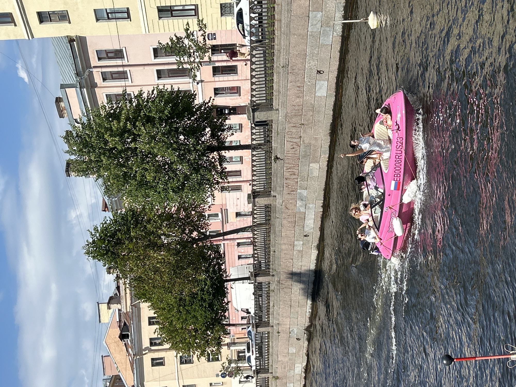 This media file shows a pink boat with several people on it, moving through a canal. The boat is a small motorboat with a "EB0008RUS23" license plate and a Russian flag on its side. The people on the boat appear to be enjoying a tour, with some looking out at the scenery and others taking pictures. The canal is lined with stone walkways and historic buildings. Trees are visible along the bank, and the sky is partly cloudy, suggesting it is daytime. The water is rippling, indicating the boat is in motion, and the overall scene conveys a relaxed and pleasant atmosphere.