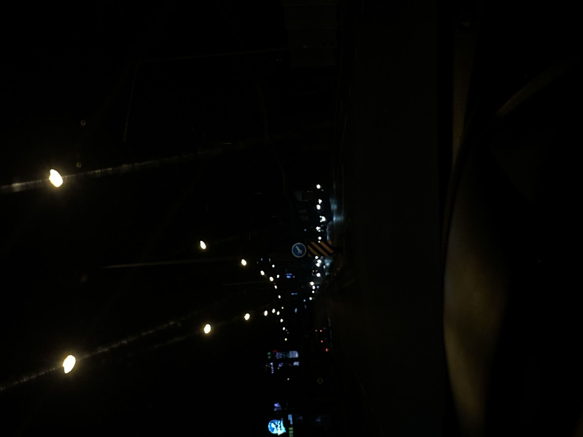 The image depicts a dark street scene in Chalong, Thailand, at night. Multiple streetlights illuminate the road, casting a yellow glow. On the left side of the street, blurred neon signs are visible, suggesting commercial activity. Several vehicles, primarily cars and motorcycles, are present on the road, their headlights and taillights creating streaks of light. A blue and white traffic sign with an arrow indicating a turn is also visible on the right side of the road. The perspective suggests the photograph was taken from inside a vehicle, likely from the driver's seat, given the view of the dashboard or hood in the foreground. The overall atmosphere is dark and atmospheric, typical of a city at night.