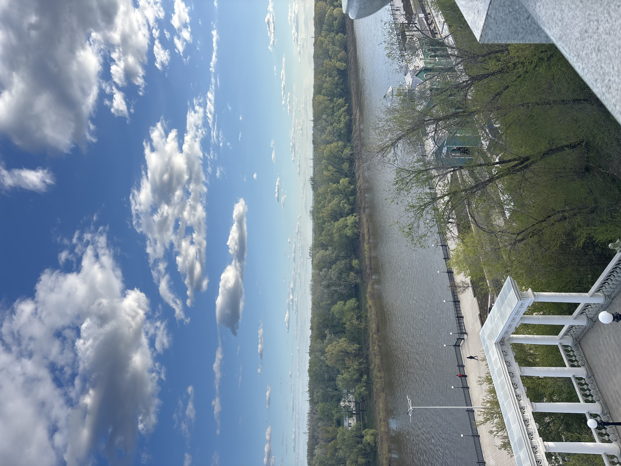 The image shows a view from a high vantage point in Orenburg, Russia, overlooking the Ural River. The sky is blue with scattered cumulus clouds. A dense forest lines the far bank of the river. The near bank has a paved walkway with decorative white colonnades and lamp posts. A few people are walking along the walkway. A white architectural structure is visible in the foreground. The overall scene depicts a bright, sunny day with natural and urban elements.