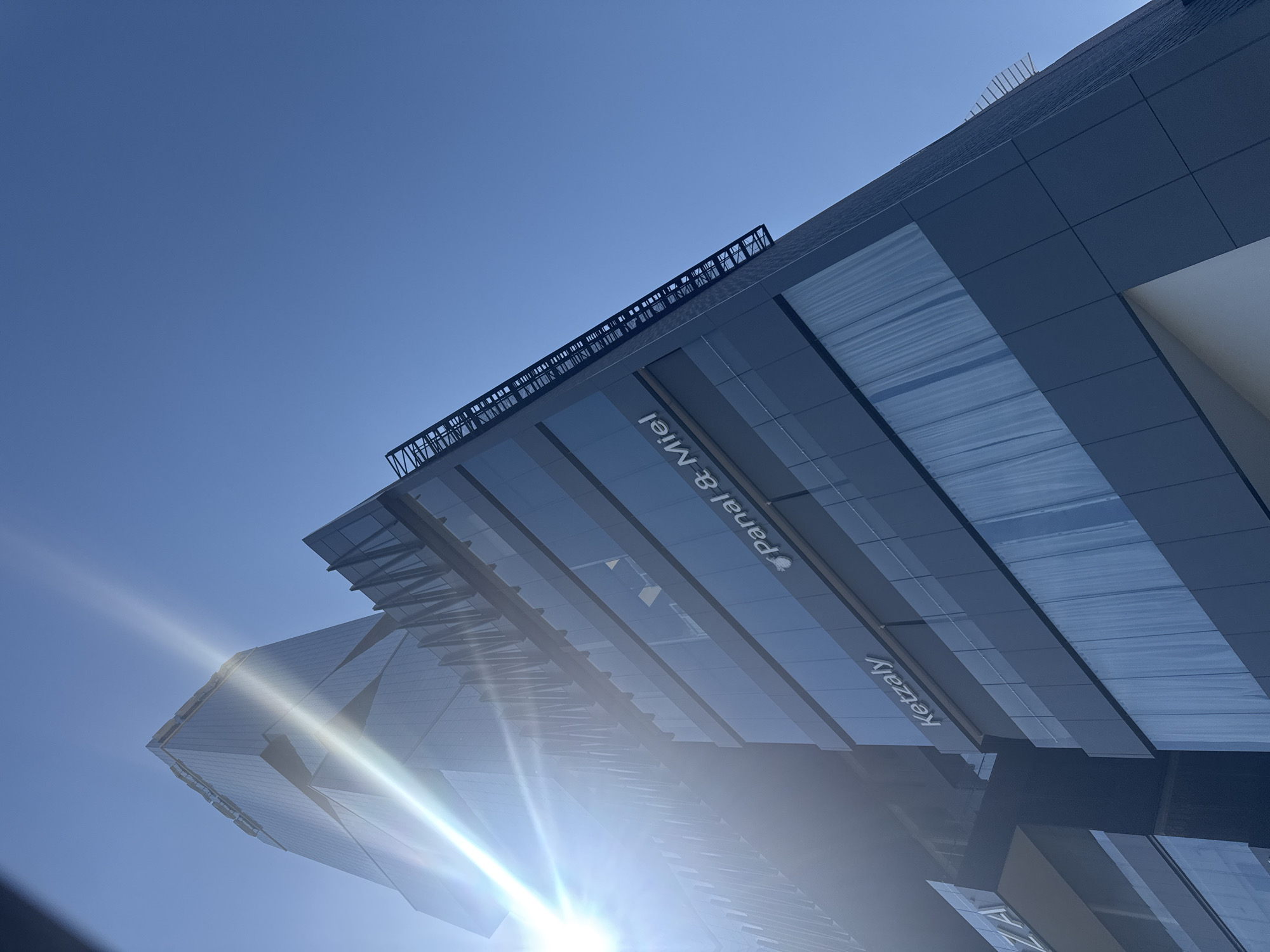 A low-angle, upward shot reveals the architectural details of a modern building in San Bernardino Tlaxcalancingo, Mexico. The building features a facade of alternating gray panels and reflective glass surfaces. A white sign bearing the name "Ketzaly" is visible, as well as the name "Panal & Miel". The sun is positioned low, creating a bright glare and lens flare that obstruct portions of the building's lower levels. The sky is a clear, deep blue. Metal latticework and other rooftop elements are partially visible along the building's edges.