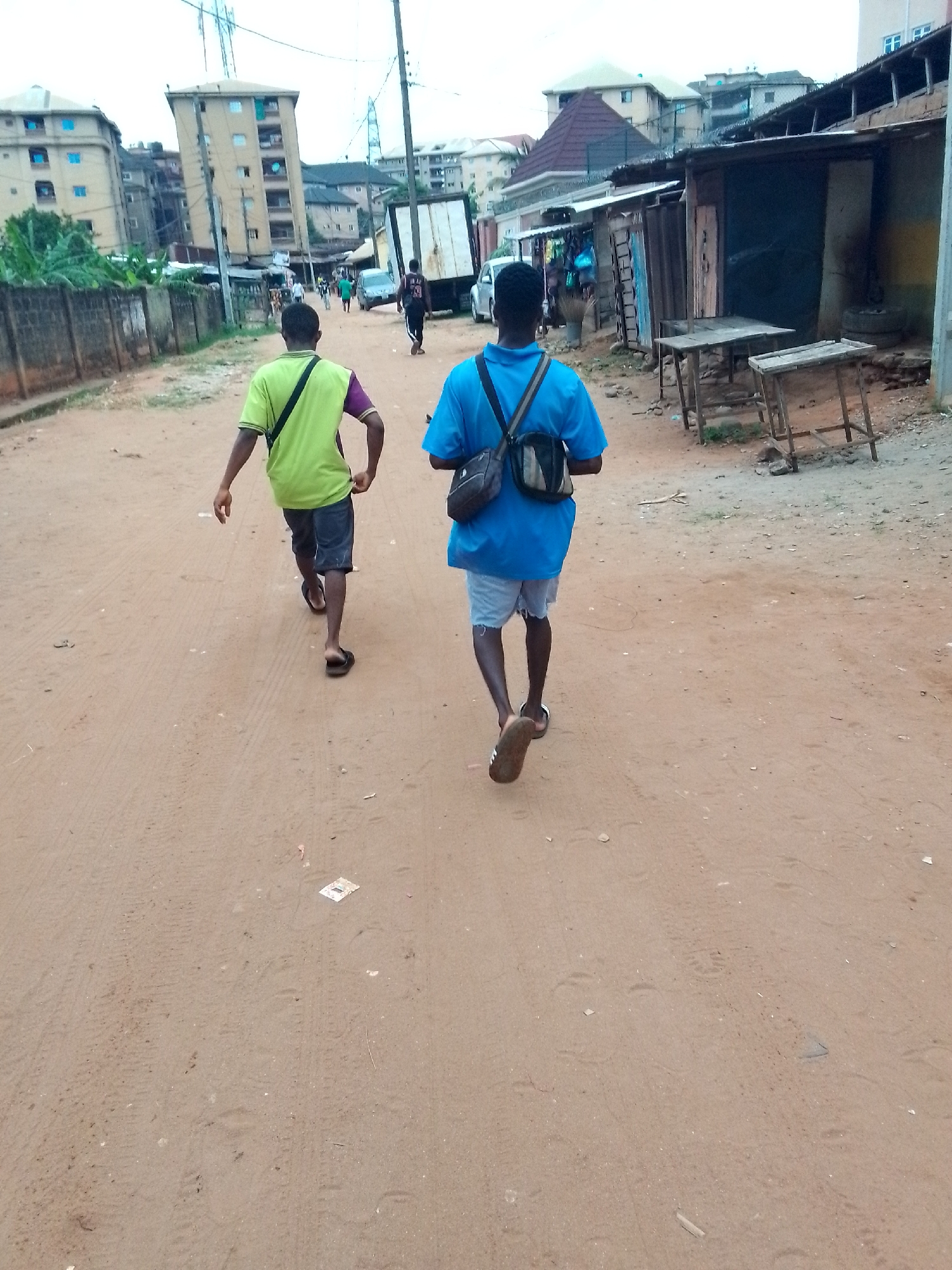 Two men walk away from the camera down a dirt road in Nkpor, Nigeria. The man on the left wears a green and purple shirt, dark shorts, and sandals. The man on the right wears a blue shirt, light shorts, sandals, and carries a shoulder bag. The road is unpaved with visible tire tracks and some litter. To the right of the road are ramshackle buildings, including open-fronted structures with tables. To the left is a grey concrete wall and buildings of varied heights. Other pedestrians are walking in the same direction. A white truck is further down the road. Power lines are visible overhead.