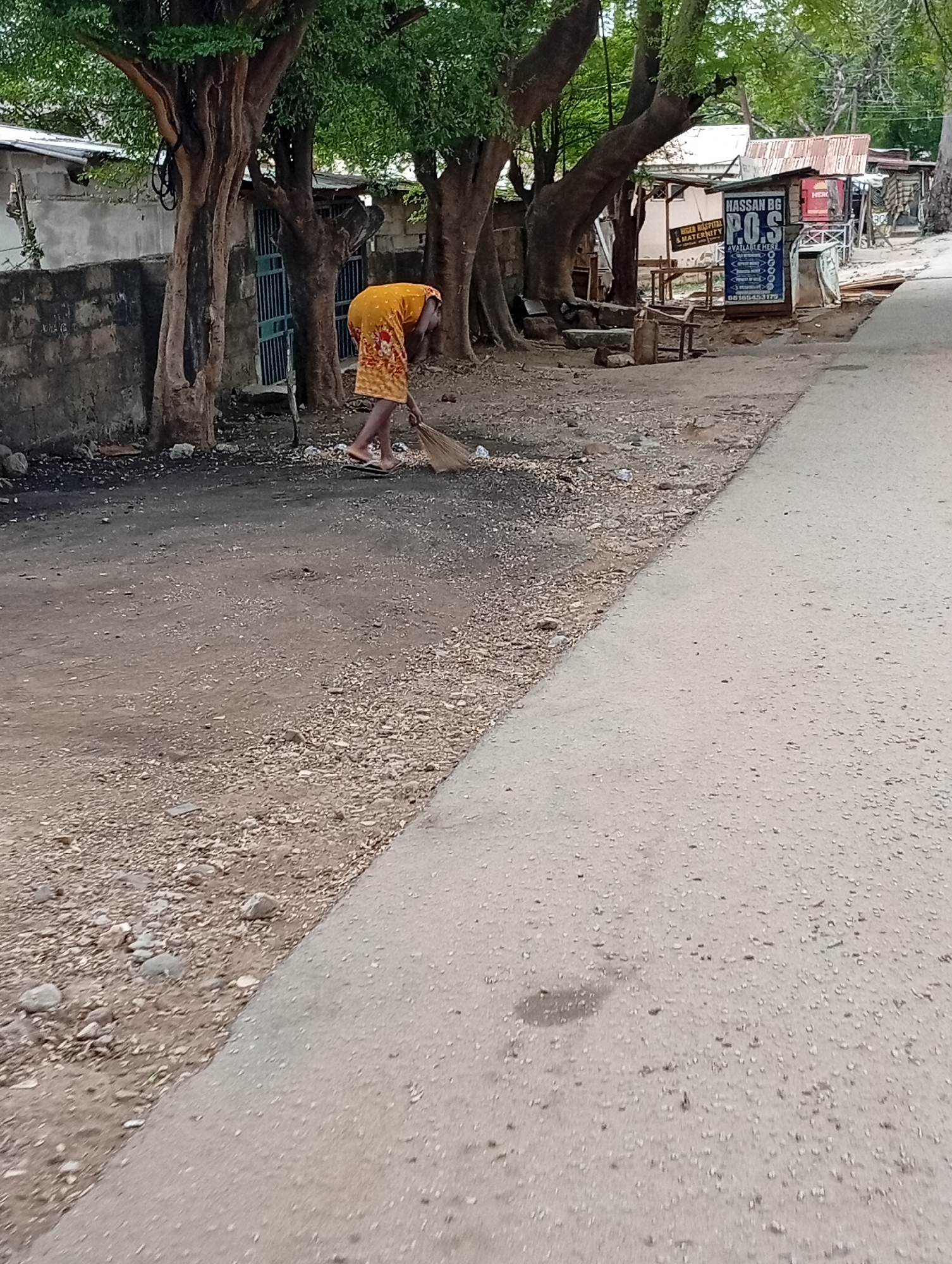 A woman in a yellow dress sweeps the roadside in Lokoja, Nigeria. The area features a mix of dirt, gravel, and paved surfaces. Trees line the roadside. A building with a sign for "HASSAN BG P.O.S" is visible in the background. Other buildings and structures are present, suggesting a commercial or residential area.