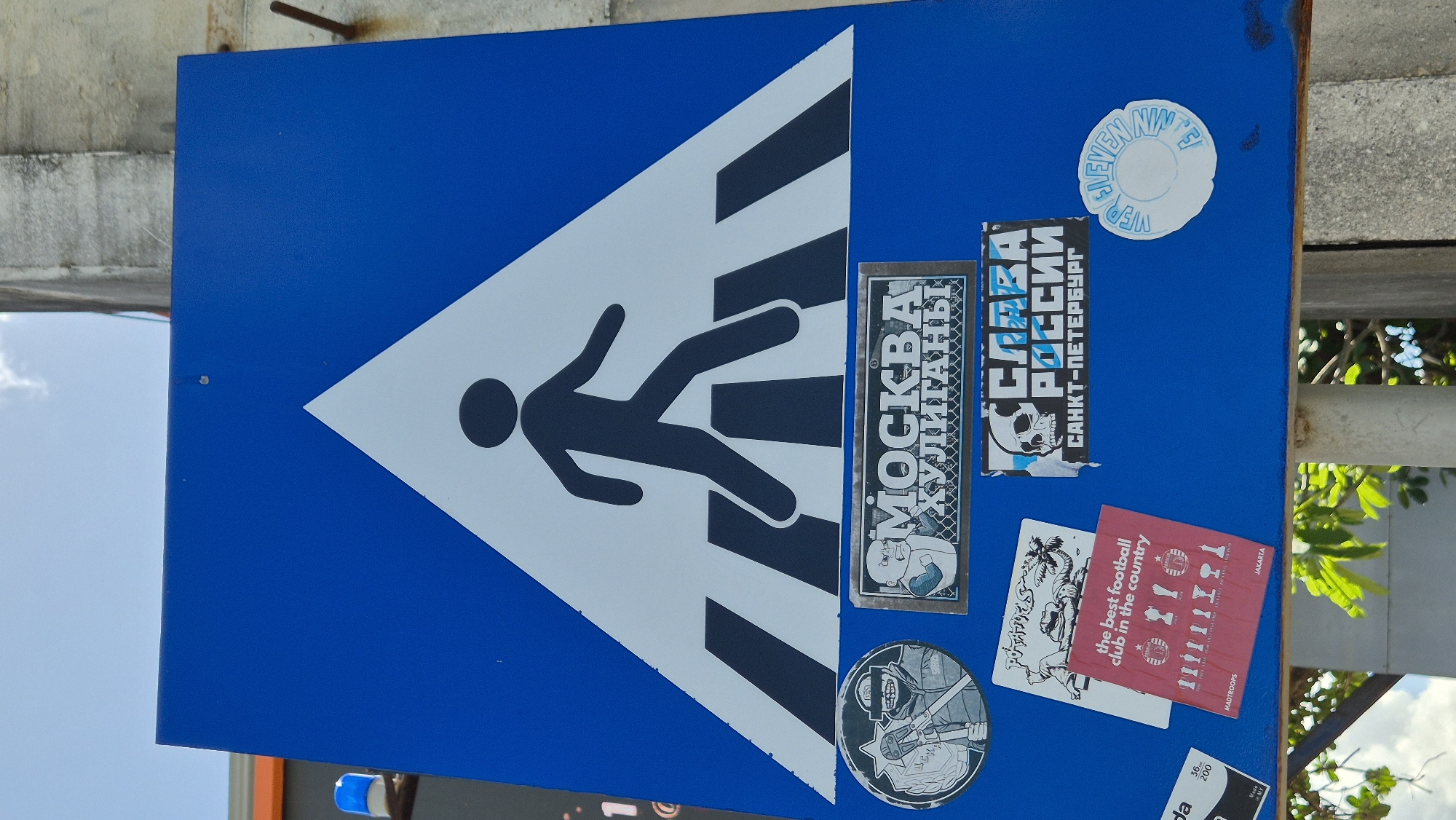 The image depicts a blue pedestrian crossing sign affixed with several stickers. The sign's primary symbol is a white triangle containing a black silhouette of a person walking across horizontal black stripes.
The stickers adhered to the sign include:
* A rectangular sticker with "МОСКВА ХУЛИГАНЫ" (Moscow Hooligans) and an image of a cartoon-like character.
* A rectangular sticker with "СЛАВА РОССИИ САНКТ-ПЕТЕРБУРГ" (Glory to Russia, Saint Petersburg), featuring a skull.
* A round sticker with lettering.
* A red card-like sticker with "the best football club in the country," featuring stick figure-like drawings of football players. It also says "JAKARTA" and "MADTROOPS".
* Other, partially visible stickers.
The sign appears to be mounted on a post or structure. The presence of a sticker referencing Jakarta suggests this image may have been taken in Kuta, Indonesia. The overall scene is somewhat urban, with greenery visible in the background.