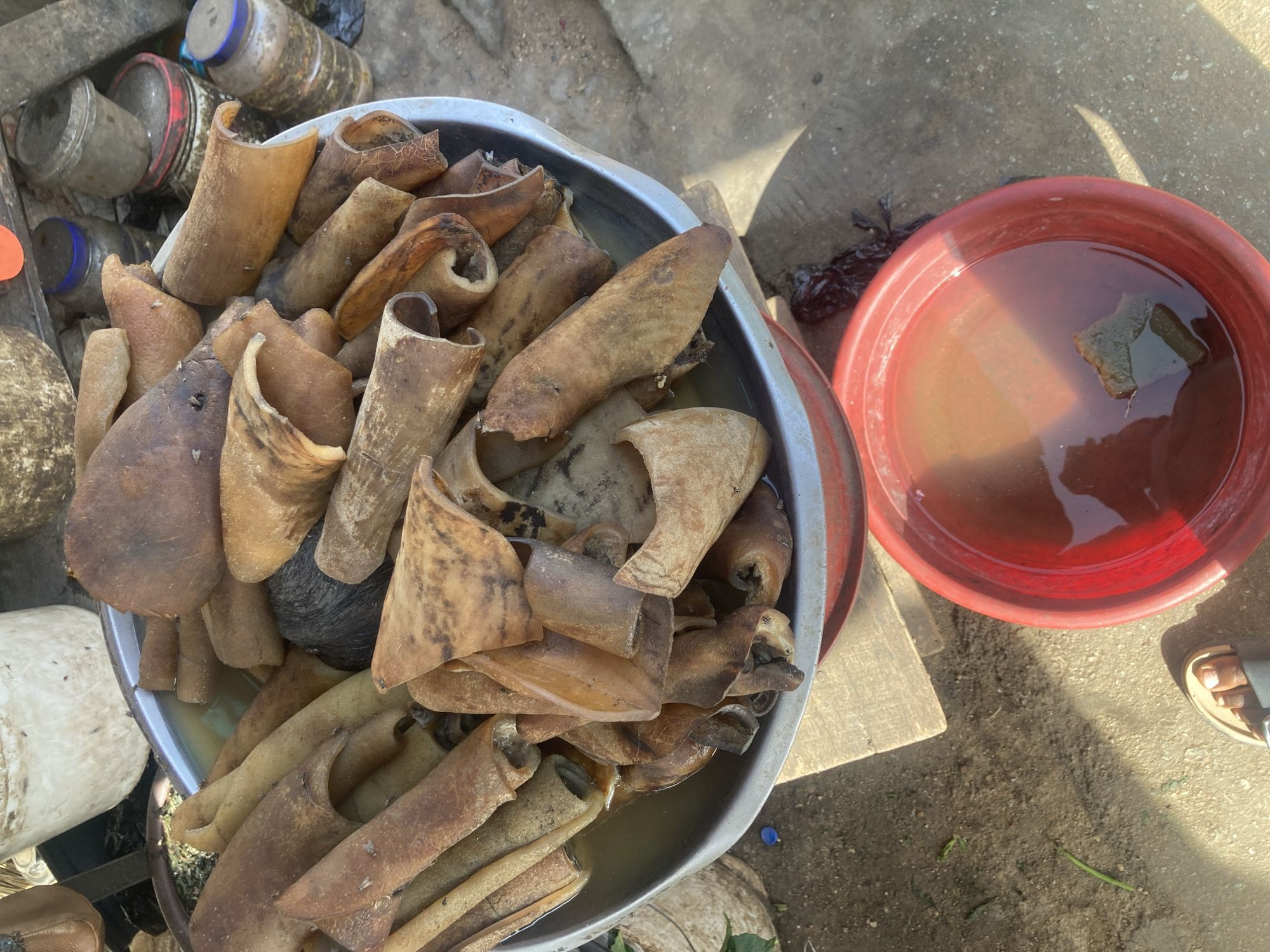 The image shows a stainless-steel bowl filled with cow skin, commonly called "Kanda" in Nigeria, submerged in water. Beside it, there's a red plastic basin also containing water with a piece of the cow skin. The background includes a collection of plastic containers and bottles. A portion of a person's foot wearing a sandal is visible. The location is presumed to be in Jos, Nigeria, based on the type of food and context. The scene appears to be a market or food preparation area.