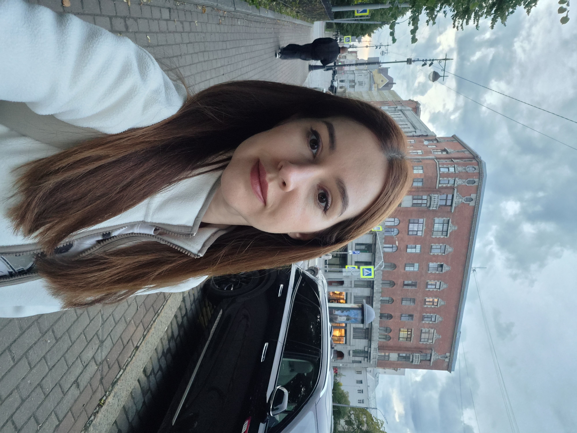 A medium shot selfie shows a woman with long, auburn hair in the foreground. She is wearing a white fleece jacket with a zipper. Behind her, a man in a dark jacket and pants walks away on a brick sidewalk. To the right, a black SUV is parked at the curb. In the background, a red brick building with architectural detailing stands under a cloudy sky. This appears to be a street scene in Vyborg, Russia.