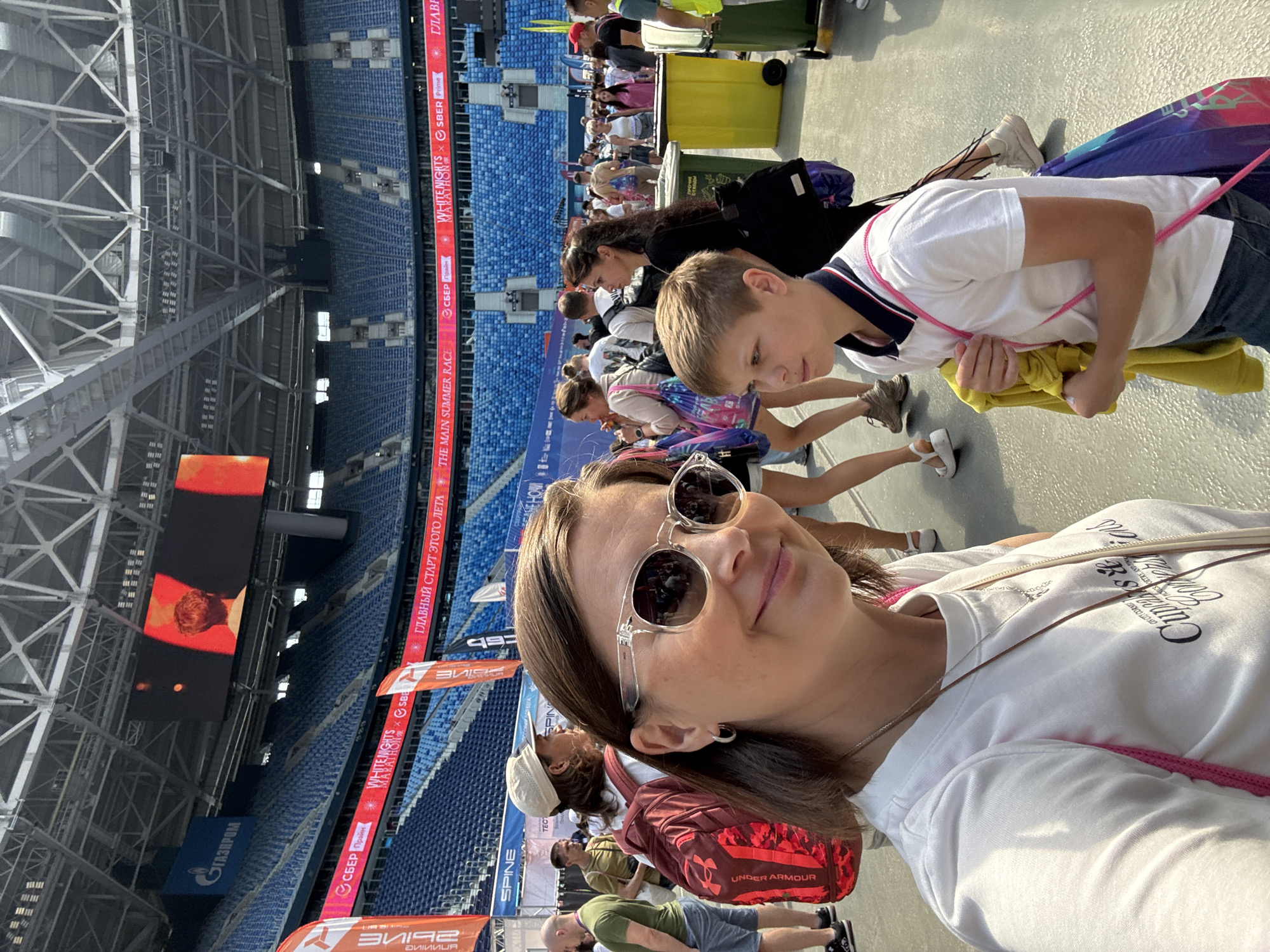 In Saint Petersburg, Russia, a woman and a young boy are taking a selfie in front of a stadium. The woman, wearing sunglasses and a white t-shirt, is in the foreground, smiling. The boy, in a white polo shirt, stands to her right. Behind them, a large stadium with blue seats is visible, along with banners advertising an event. People are milling about in the background, indicating a public gathering or sporting event.