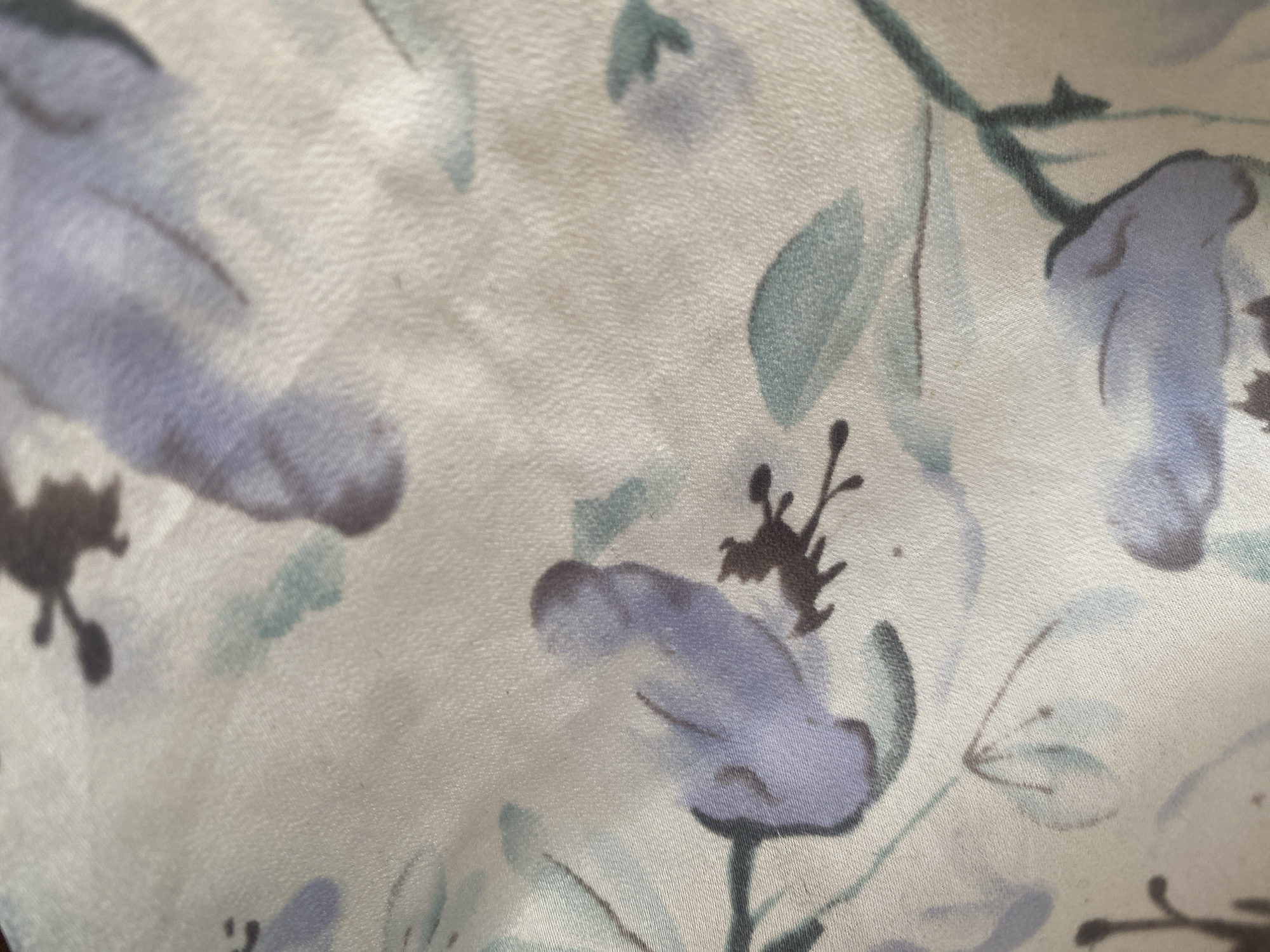 This media file is a close-up, low-angle shot of a light-colored fabric with a floral pattern. The fabric appears to be textured, with visible weave and slight sheen, suggesting it might be silk or a similar material. The pattern features abstract depictions of flowers and leaves in muted shades of purple, blue, and green, with some darker accents. The overall impression is one of softness and a delicate aesthetic. There are no people, explicit objects, or a discernible scene depicted; the focus is solely on the texture and pattern of the fabric. The lighting is soft, and there are no strong shadows, indicating an indoor setting or diffused natural light. The image lacks any contextual cues related to time of day, weather, or specific location beyond the implied presence of fabric, which is not tied to Dumnga, Nigeria. No text is visible in the image.