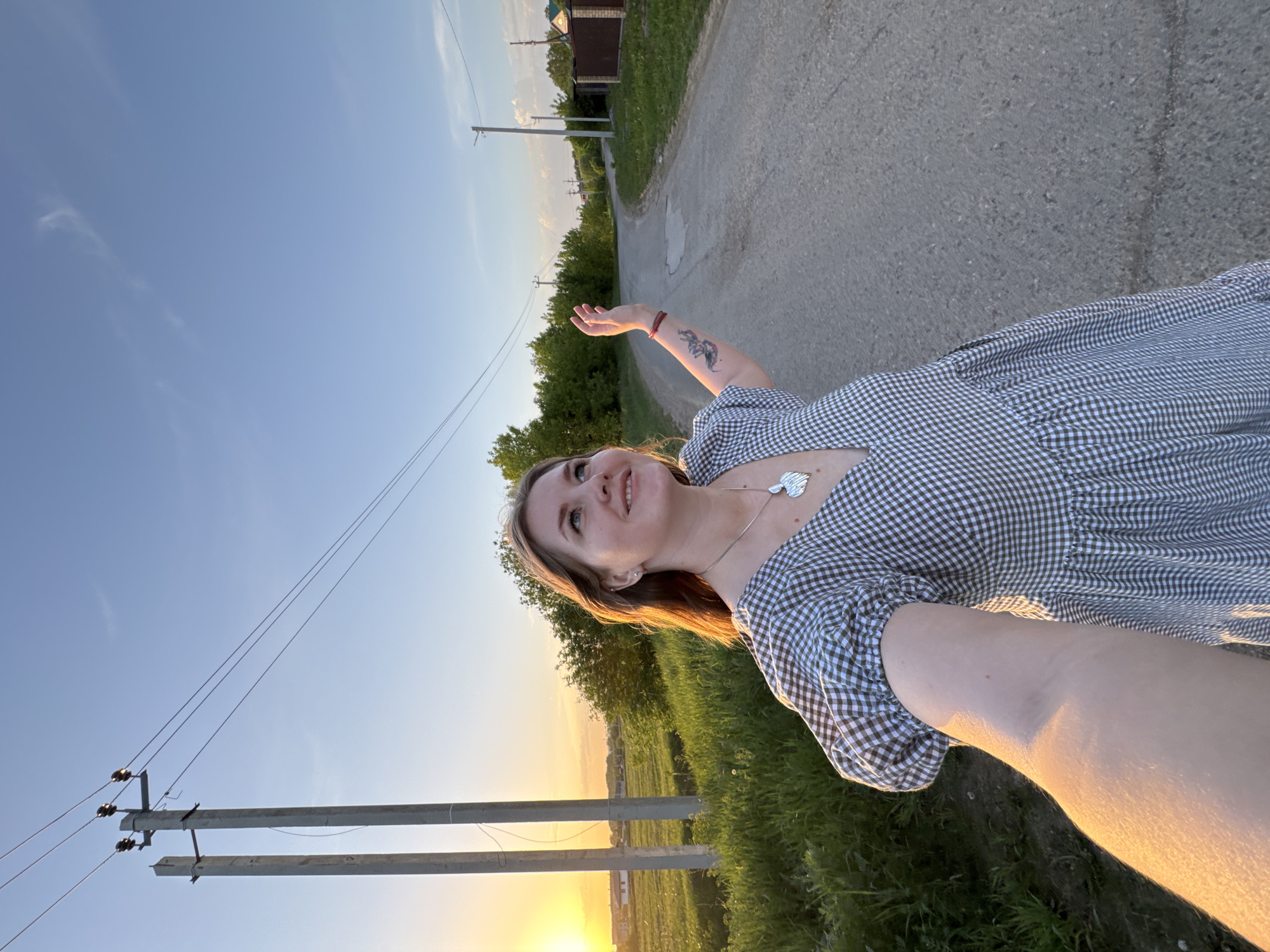 A photograph depicts a woman standing on an asphalt road in Горный, Russia. The road, with a patch repair visible, stretches into the distance where a building with a dark roof is situated. The woman, dressed in a checkered dress, has her arm raised. She also has a tattoo on her arm and a silver pendant. In the background, green vegetation is abundant on either side of the road, with electrical poles and wires running alongside. A sunset is visible on the horizon, casting a warm glow on the scene.