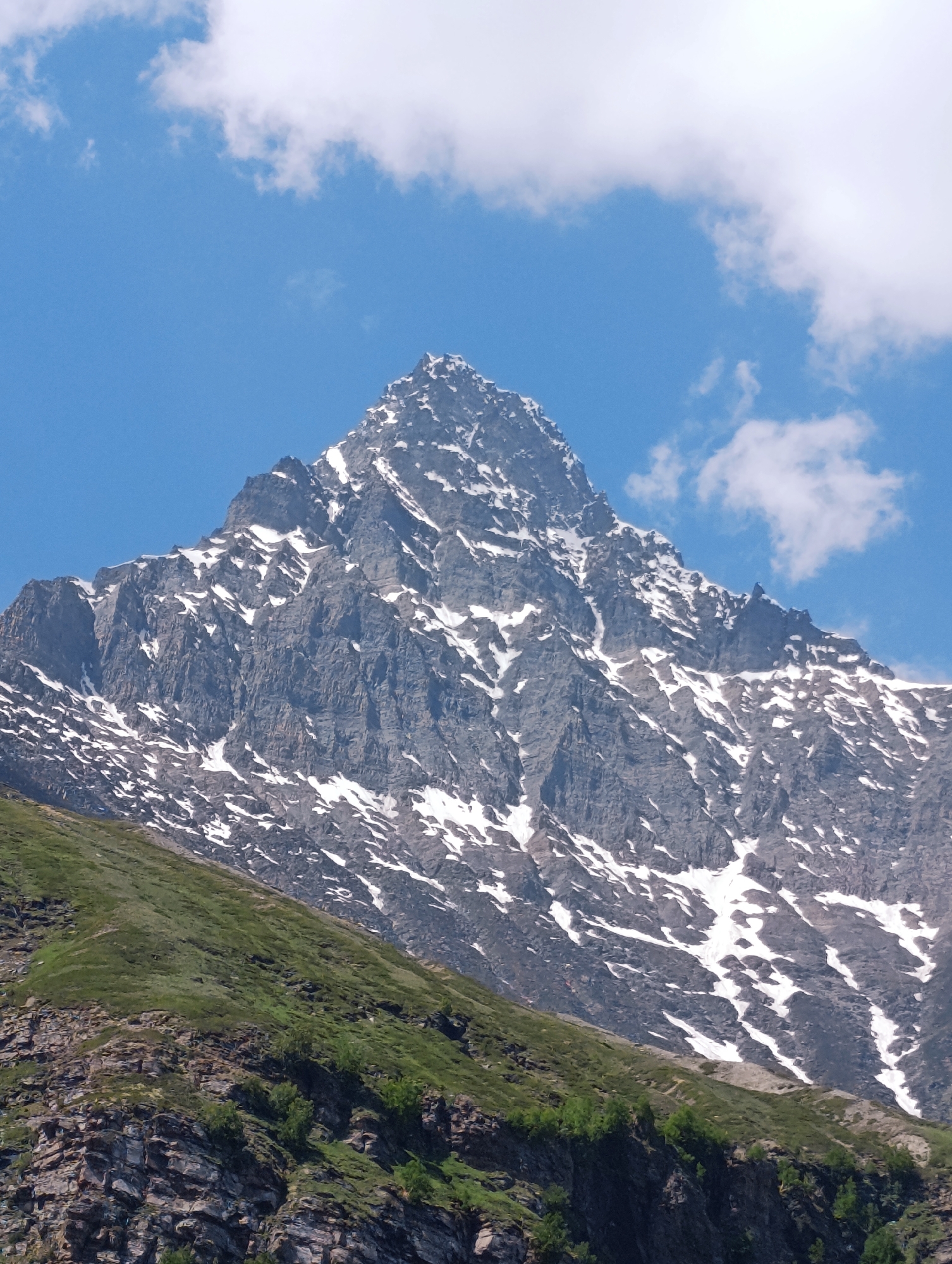 A vertical, low-angle, eye-level shot captures a majestic snow-capped mountain peak under a bright blue sky with scattered white clouds. The mountain itself is rugged and rocky, with dramatic slopes and jagged formations. Streaks of white snow adorn its upper reaches, contrasting with the dark gray rock. The lower slopes transition into verdant green, with patches of rocks and a few sparse trees visible. The image is taken from the foothills, looking upwards at the imposing mountain. The scene evokes a sense of natural grandeur and the raw beauty of the Himalayas. The specific location is not identifiable from the image alone, but it is a mountainous landscape that could be found in various regions of India.