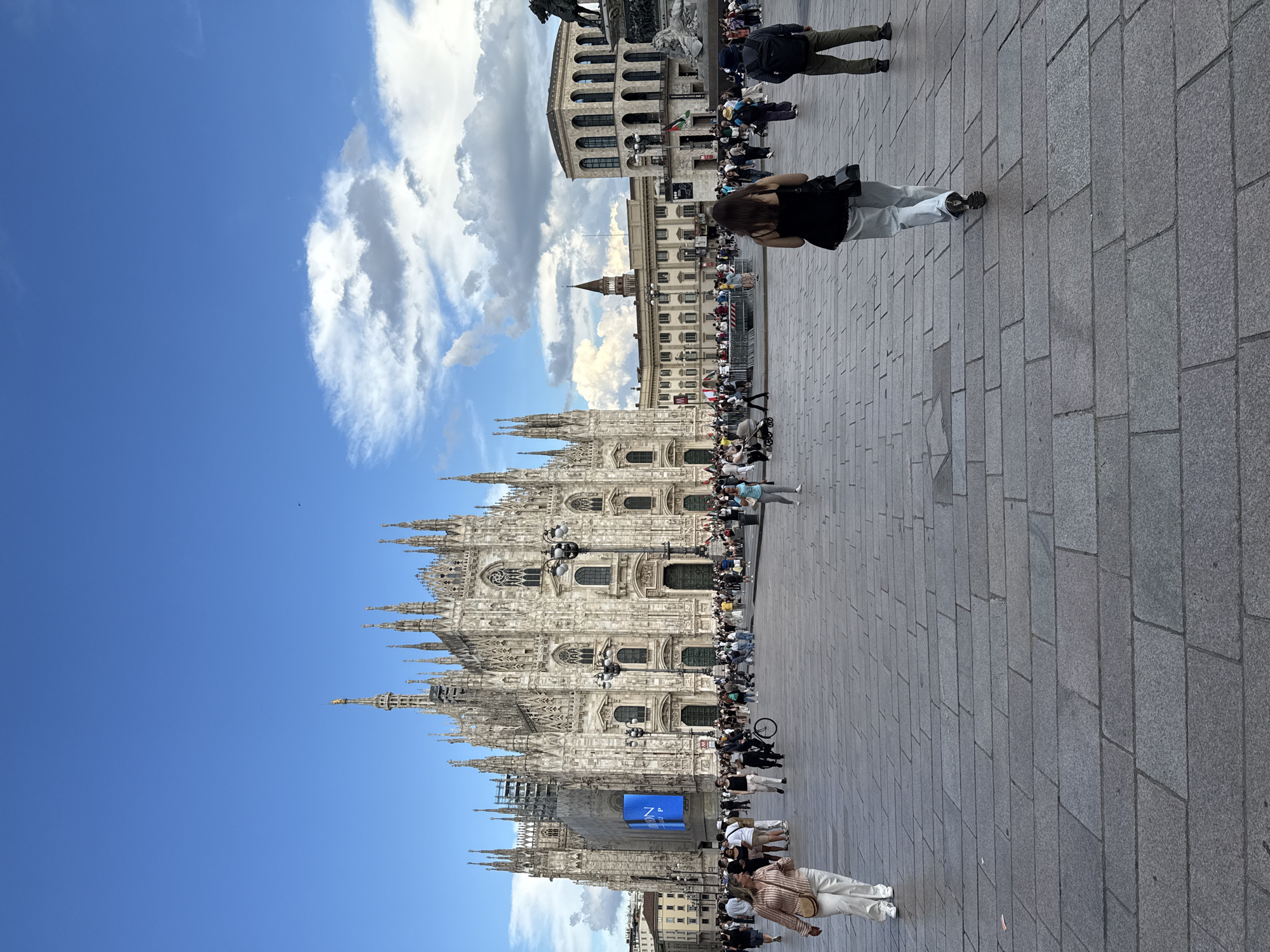 The image depicts Piazza del Duomo in Milan, Italy, under a bright blue sky with scattered white cumulus clouds. The central feature is the Duomo di Milano, a large Gothic cathedral with intricate white marble facades, numerous spires, and detailed sculptures. Scaffolding is visible on the left side of the cathedral, partially obscured by a prominent blue banner displaying white text.
The piazza foreground is paved with grey rectangular stone slabs. A significant number of people are distributed across the mid-ground, engaging in various activities such as walking, observing, and gathering. In the immediate foreground on the left, a woman with light-colored hair, wearing a striped shirt, white pants, and a light-colored shoulder bag, walks towards the left side of the frame. On the right foreground, a woman with dark hair, wearing a black top and light-colored pants, walks away from the camera, followed by a man with a dark backpack.
In the mid-ground crowd, individuals are dressed in casual attire. Barriers are present in the central-left area, and flags, including one resembling the Palestinian flag, are visible within a gathering on the right side of the piazza. Streetlights with multiple lamps are positioned throughout the square. Other historical buildings, including one with prominent arched windows and another with a smaller tower, are visible surrounding the piazza.
