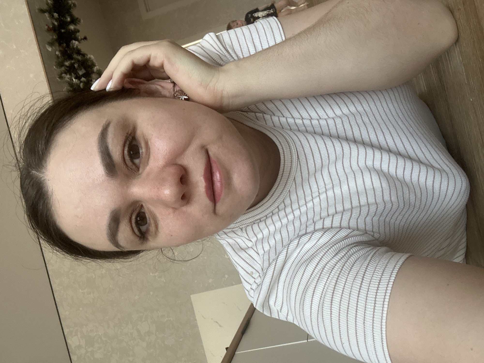 A young adult woman is captured in a selfie taken indoors in Chaikovsky, Russia. The image is oriented vertically, suggesting she is lying down or leaning back, looking directly into the camera with a gentle, slight smile. She has dark hair, dark brown eyes, and full eyebrows. Her skin appears clear and well-hydrated, and her lips have a natural pink tone. She is wearing a light-colored (white or off-white) top with thin, vertical brown stripes. Her right hand, adorned with a ring on her ring finger and white-painted fingernails, is resting near her head. In the upper left background, a glimpse of a decorative garland or similar plant with white accents is visible. The setting appears to be a home interior, featuring light-colored wooden surfaces and walls. A faint, blurred reflection of another individual, possibly a child, can be seen in the background on the right side of the frame. The lighting indicates it is daytime. There is no visible text.