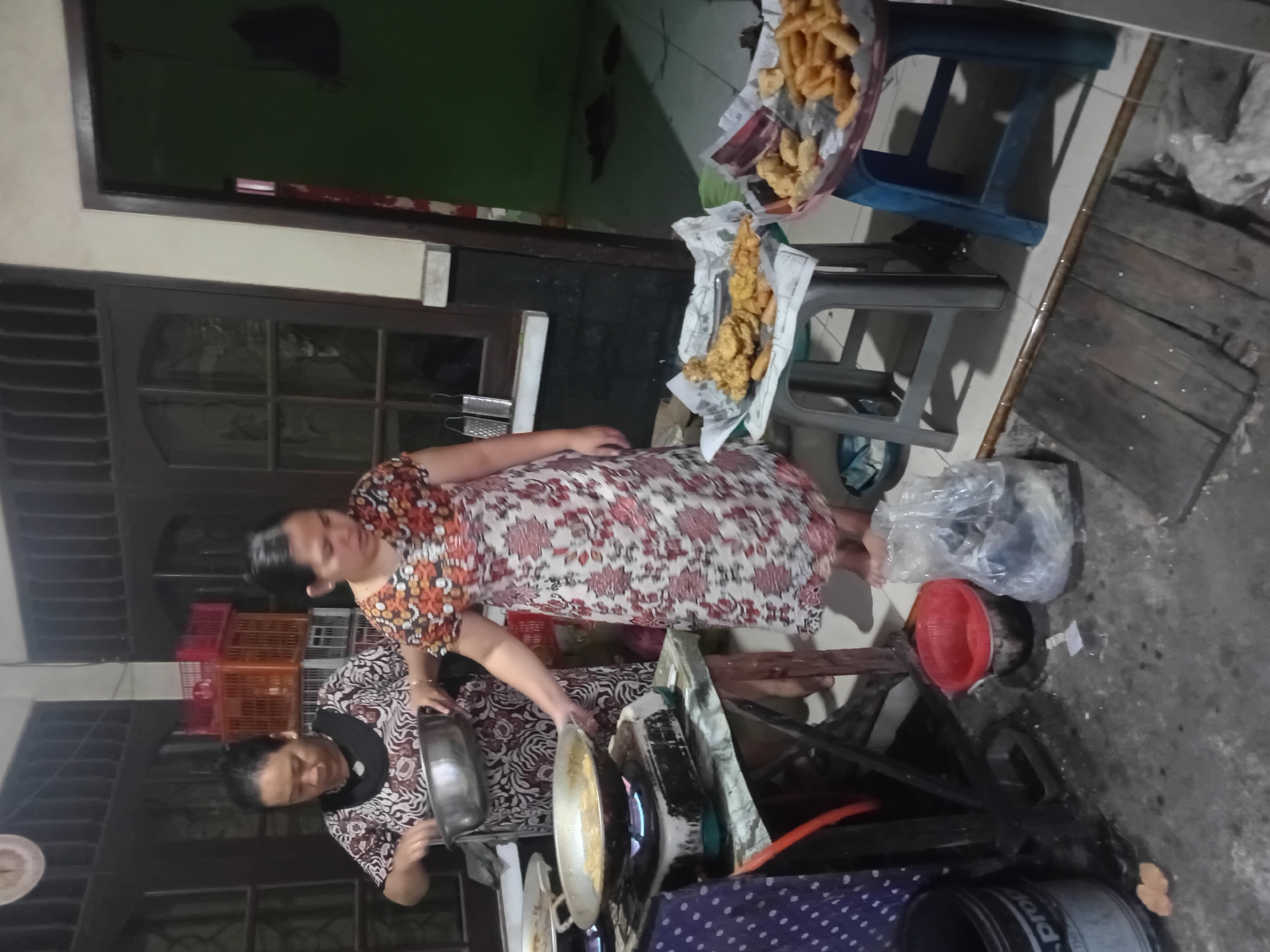 In Besito, Indonesia, two women are engaged in food preparation. The woman on the left, wearing a patterned dress, is holding a metal bowl and appears to be pouring something into a frying pan on a gas stove. The woman on the right, in a floral dress, is standing and looking at the pan. The scene takes place in a dimly lit indoor space with tiled floors and dark wooden doors or window frames. Various cooking implements and containers, including red buckets and plastic bags filled with food, are visible. In the foreground, two plastic stools hold trays piled high with fried snacks, likely for sale or consumption. The overall impression is one of domestic food preparation, possibly for a local market or event.