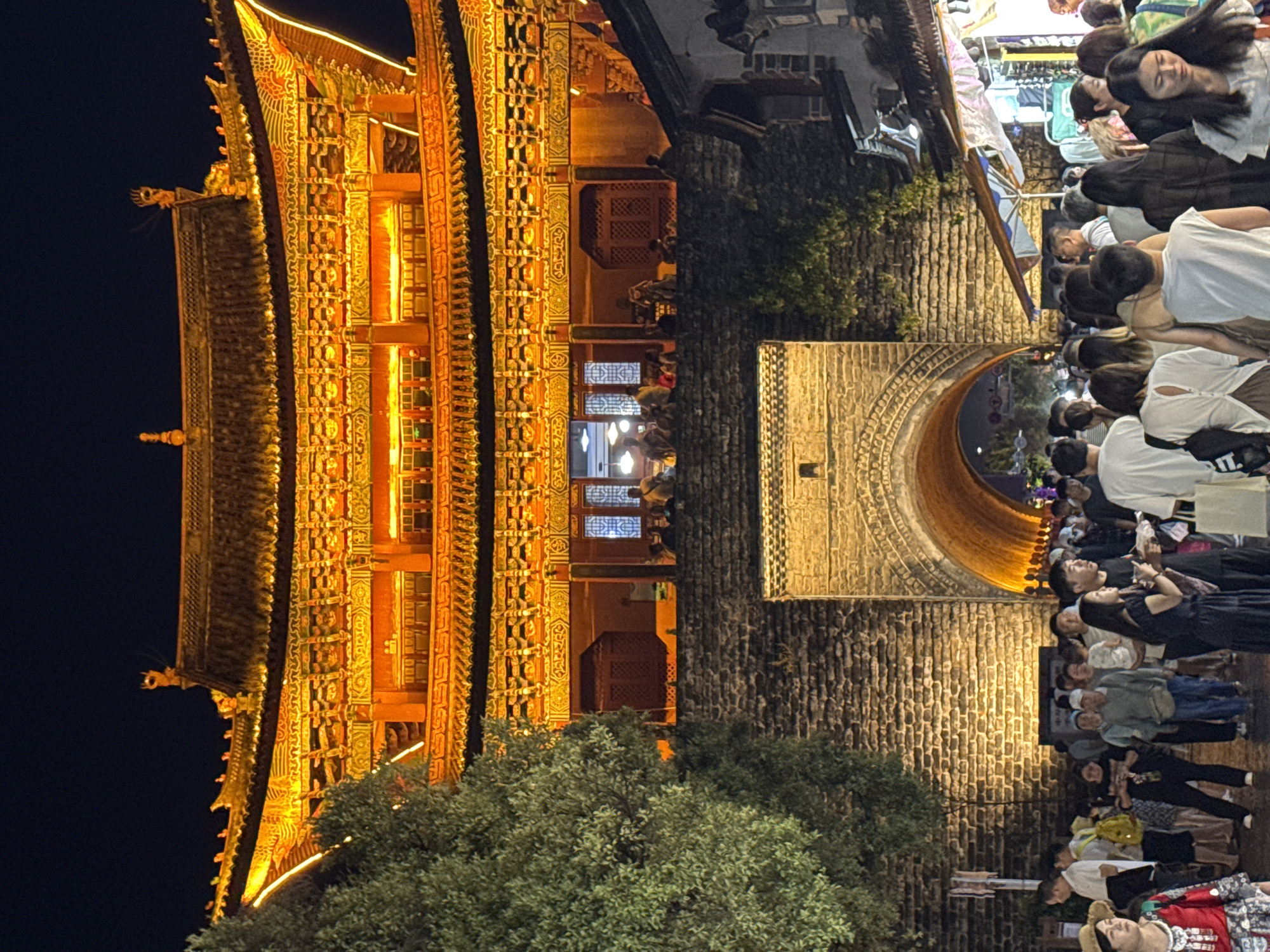 This is a nighttime outdoor vertical full shot of a pedestrian area in Dali City, China, featuring an illuminated ancient gate structure and a crowd of people. The gate is constructed of rough stone blocks and features a large arched entryway that glows with orange light from within. Above the stone gate is a multi-tiered, ornate building with a brightly lit, orange-hued roof and intricate detailing. Red and yellow lights are strung along the eaves of the building. A large, leafy green tree is visible to the left of the gate. A dense crowd of people, dressed in casual clothing, is walking in front of and through the gate. Some individuals are carrying bags. The ground appears to be wet, suggesting it may have rained. The overall atmosphere is lively and festive, with the bright lights illuminating the scene and highlighting the architectural features.