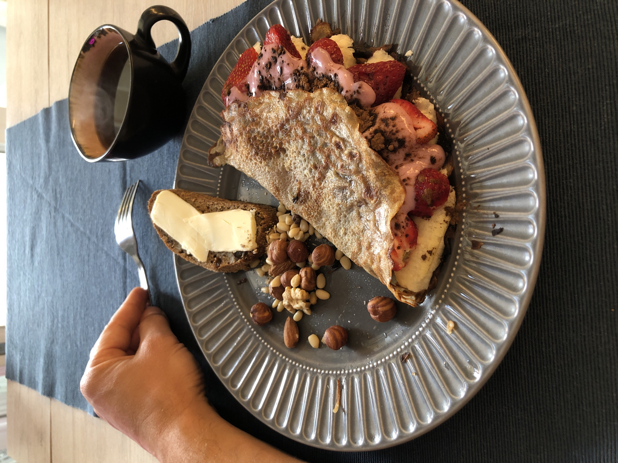 A close-up, overhead shot shows a breakfast spread on a dark blue placemat atop a wooden table. A person's left hand is visible in the upper left corner, holding a fork poised over a slice of dark bread topped with butter. To the right, a gray plate holds a folded crepe filled with strawberries and a pink sauce, accompanied by a scattering of nuts and pine nuts. A black mug filled with steaming coffee sits to the upper right of the plate. The scene depicts a meal at home.