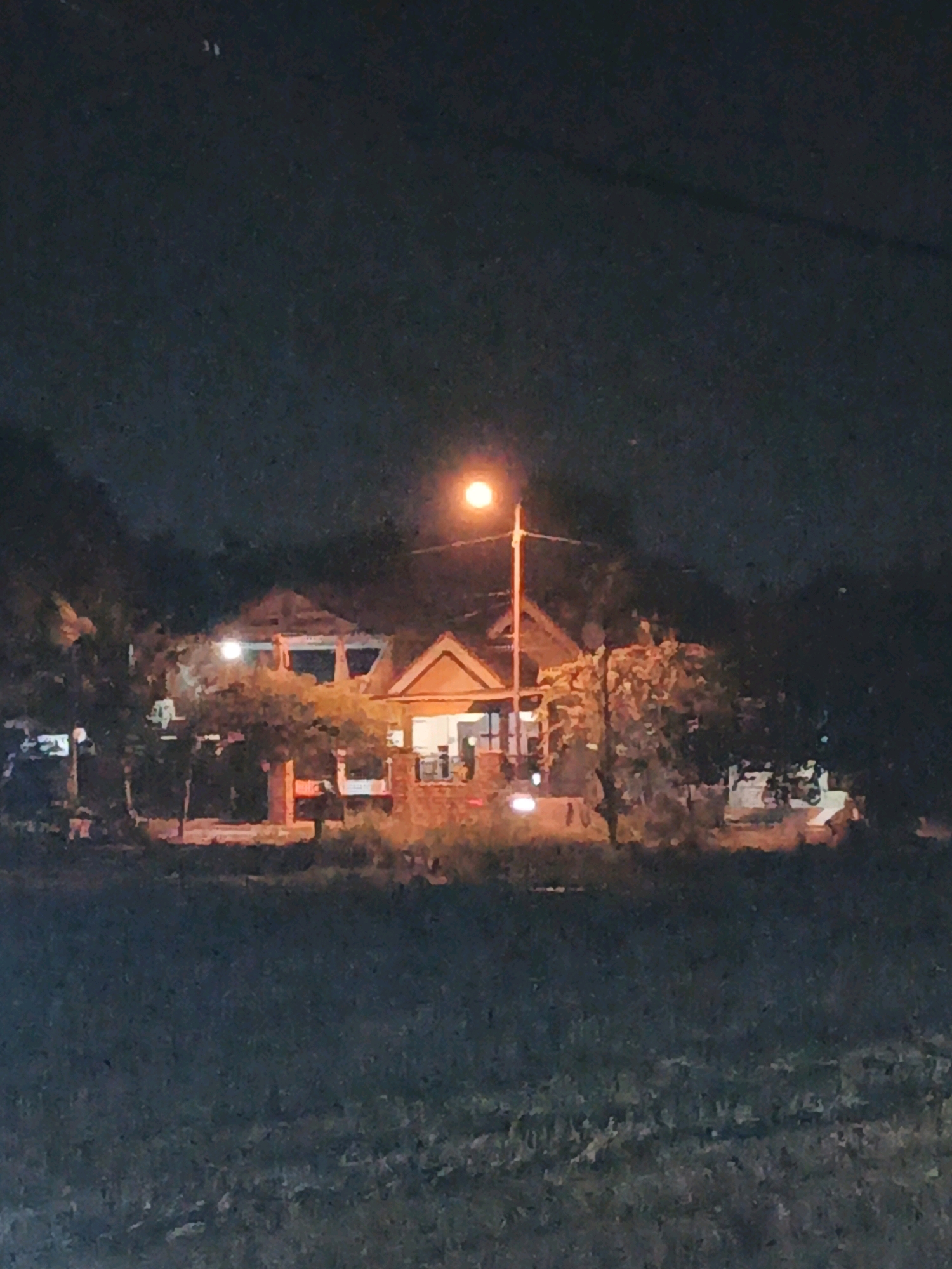 A nighttime shot taken from a distance shows a house with lights on, surrounded by trees. A streetlamp illuminates the scene, casting an orange glow on the building and vegetation. The image is grainy and out of focus, making it difficult to discern specific details. The foreground appears to be a grassy area.