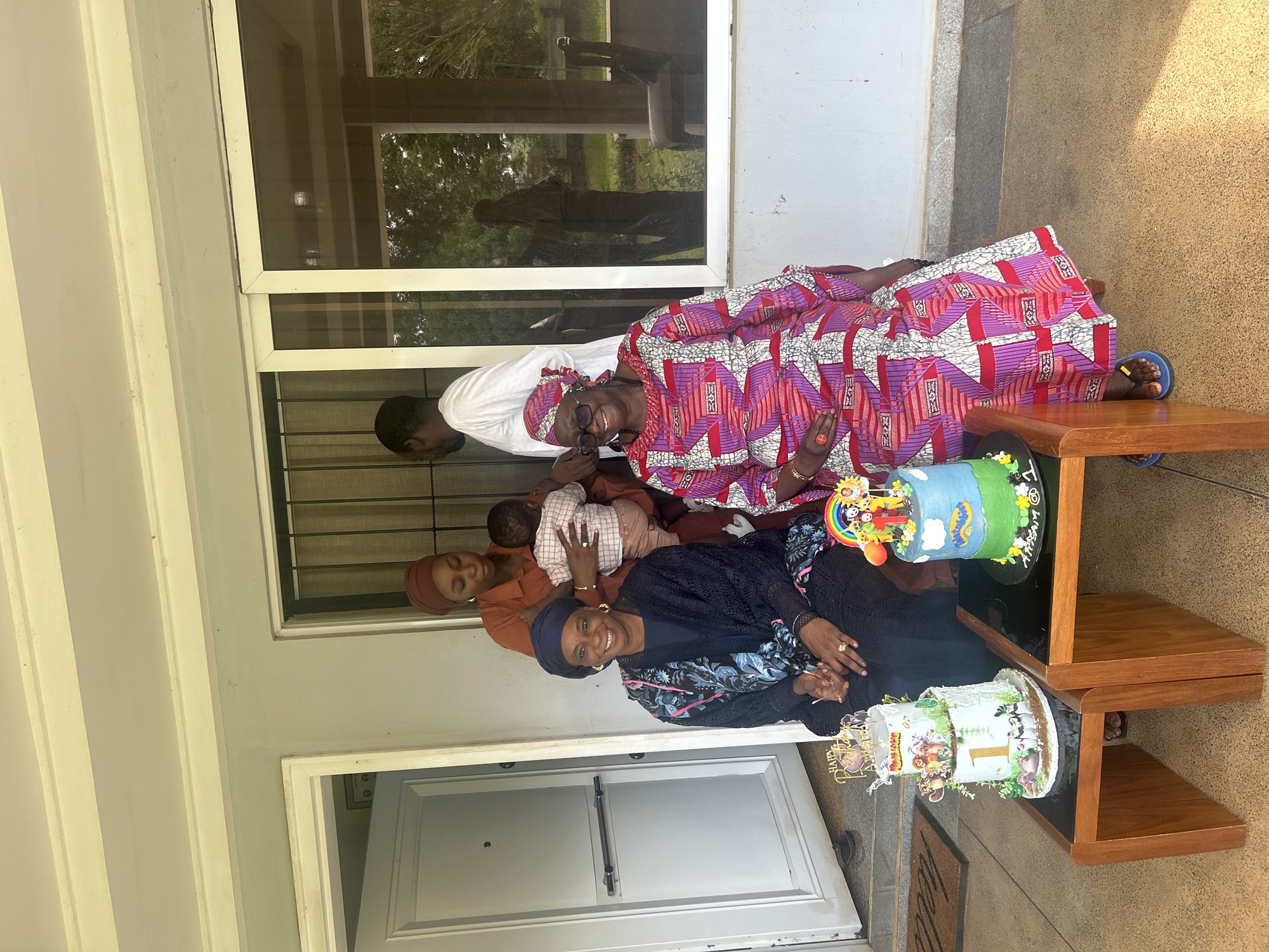 In Dutsen Kato, Nigeria, at a building with a covered porch, four people are gathered. Two women are seated in the foreground. The woman on the left wears a dark blue outfit and headscarf and smiles. The woman on the right wears a vibrant, patterned dress and glasses and smiles broadly. In front of them, on two wooden tables with black tops, are two tiered cakes. The cake on the left is white with a gold number 1 and cartoon animal figures. The cake on the right is blue and green with "Twistables" written on it and cartoon figures. Behind the seated women, a third woman in an orange headscarf holds a baby. A man in a white shirt stands behind her. The background shows a door on the left and a large window with reflections of trees and furniture on the right.