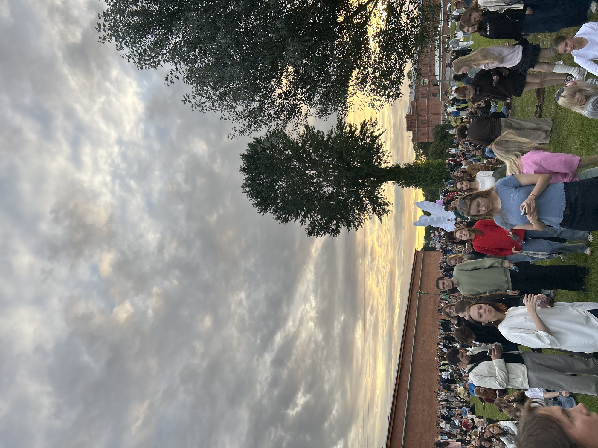 The image depicts a large outdoor gathering of people on a grassy area in front of a brick building under a cloudy sky at dusk. The gathering appears to be in a park or open space. In the background, a large brick building with multiple windows and a red-tiled roof is visible, along with trees. There is a white, abstract sculpture in the middle-ground. The sky is filled with dramatic clouds, with streaks of orange and yellow suggesting the setting sun. The people are dressed in casual clothing and many are holding phones. Some are standing, while others are sitting on the grass. The scene suggests a public event or festival taking place in a historical urban setting, likely the Peter and Paul Fortress in Saint Petersburg, Russia, given the architectural style of the building and its common use as an outdoor venue.