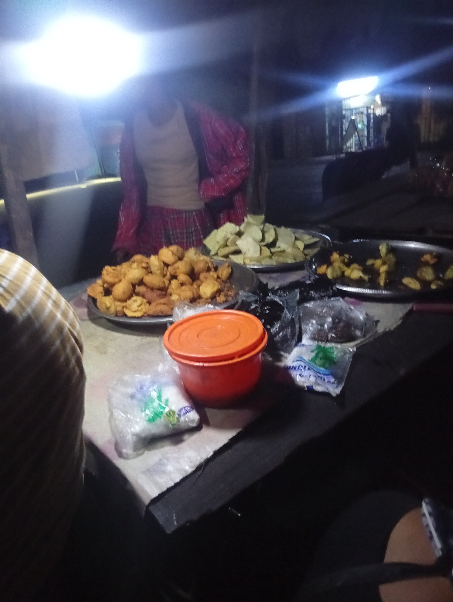 The image depicts a dimly lit outdoor food stall, likely in Nkpor, Nigeria, at night. A person, whose face is obscured, is visible behind a counter covered with food items. The person is wearing a red and white checkered shirt and a light-colored undershirt.
On the counter, there are several platters of food. One platter contains numerous golden-brown fried items, possibly fritters or dumplings. Another platter holds sliced, light-green food, which could be plantains or yams. A third platter contains smaller, darker fried pieces. An orange plastic container with a lid sits prominently on the counter, along with several plastic bags containing food items. One bag, partially transparent, shows green sprigs within, possibly herbs or spices.
The stall appears to be made of rudimentary materials, with exposed wooden posts and a corrugated iron roof. The background is dark, with hints of a structure and some lights suggesting other stalls or buildings further away. The overall scene suggests a typical nocturnal street food vending operation in an urban Nigerian setting.