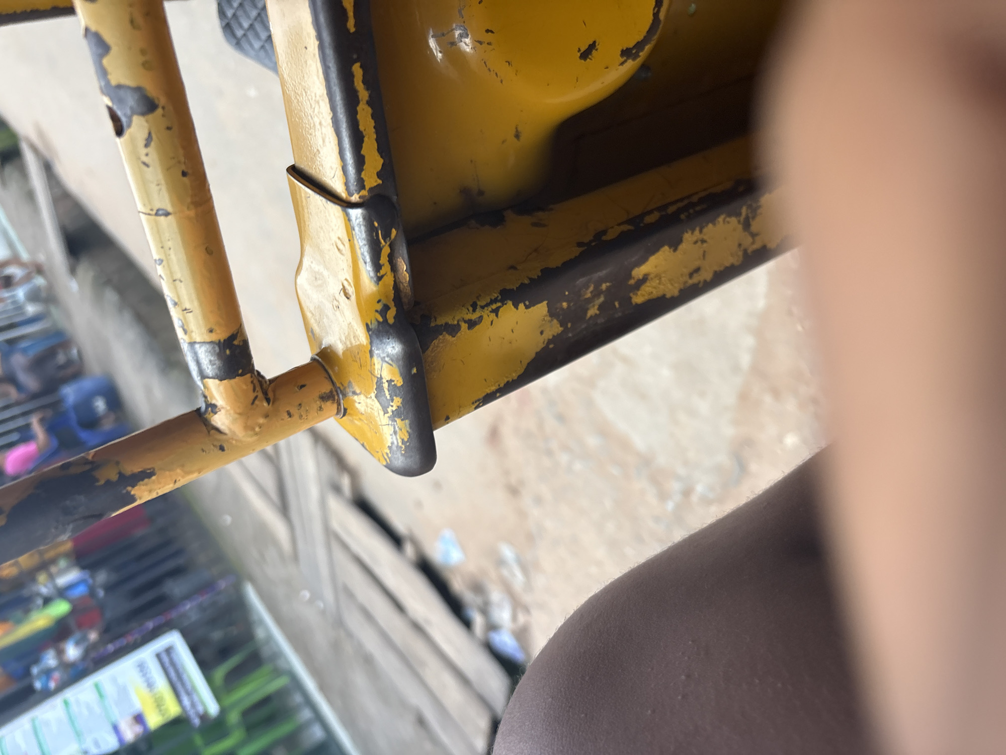A close-up, slightly blurred shot shows a section of a yellow metal frame with peeling black and yellow paint. The frame appears to be part of a vehicle, possibly a motorcycle or tricycle, common for transportation in Onitsha, Nigeria. In the background, wooden planks are visible, suggesting a path or structure made of wood. Beyond the planks, the ground is a mixture of dirt and debris, with some plastic waste scattered. Further in the background, a sign with text is partially visible on the left, and a person in a pink shirt and blue shorts is seated, indicating a public or commercial area. The overall scene depicts a somewhat weathered and utilitarian environment, typical of street scenes in some parts of Onitsha.