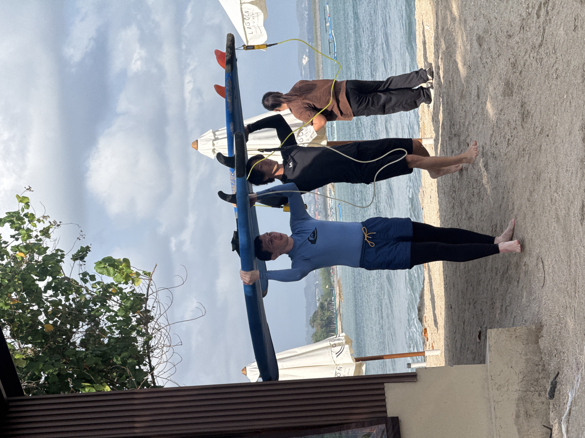 Three people are on a sandy beach near the ocean in Kuta, Indonesia. Two men are carrying a blue surfboard overhead, with the one on the left wearing a blue long-sleeved rash guard and blue shorts over black leggings, and the one in the middle wearing a black wetsuit and black shorts. A woman in a brown long-sleeved shirt and black pants stands next to them, appearing to be assisting. In the background, there are beach umbrellas, the ocean, and a distant coastline with trees. The sky is overcast with clouds.