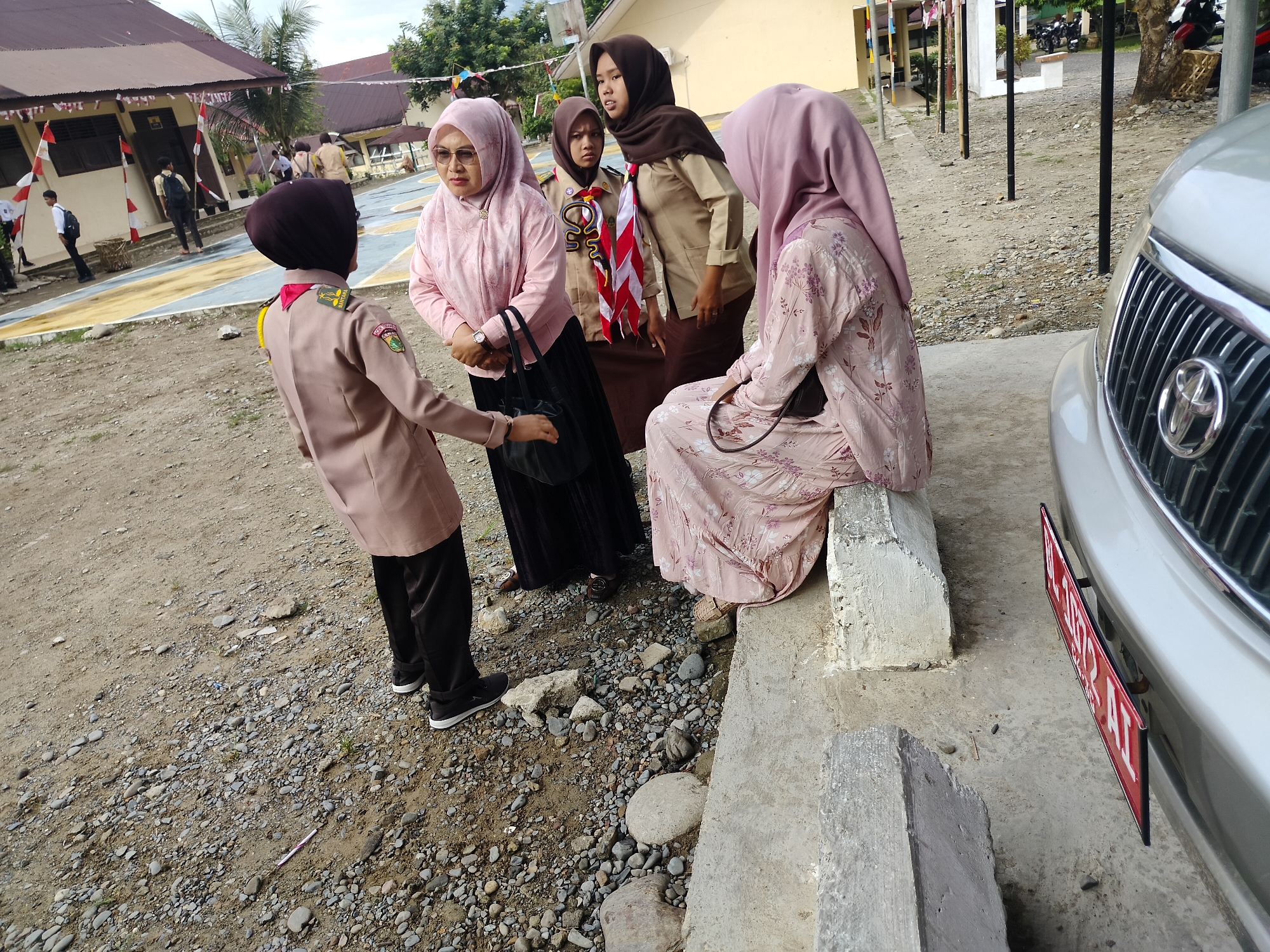 In Lawebekung, Indonesia, a group of four individuals, three women wearing hijabs and one young person in a scout uniform, are gathered outdoors near a building with red and white flags. The woman on the far left is wearing a light pink patterned top and a pink hijab. She is looking down and holding a black bag. To her right, another woman in a similar pink top and hijab, with glasses, is speaking with her. Behind them, a young person in a tan scout uniform with a red and white neckerchief stands between two adult women. The woman on the far right is seated, wearing a light pink patterned dress and hijab, facing the other individuals. In the foreground, a silver car with a red license plate is partially visible, parked on a concrete surface. The ground is covered with gravel and dirt. The background shows a building with a brown roof and windows, and some trees.
