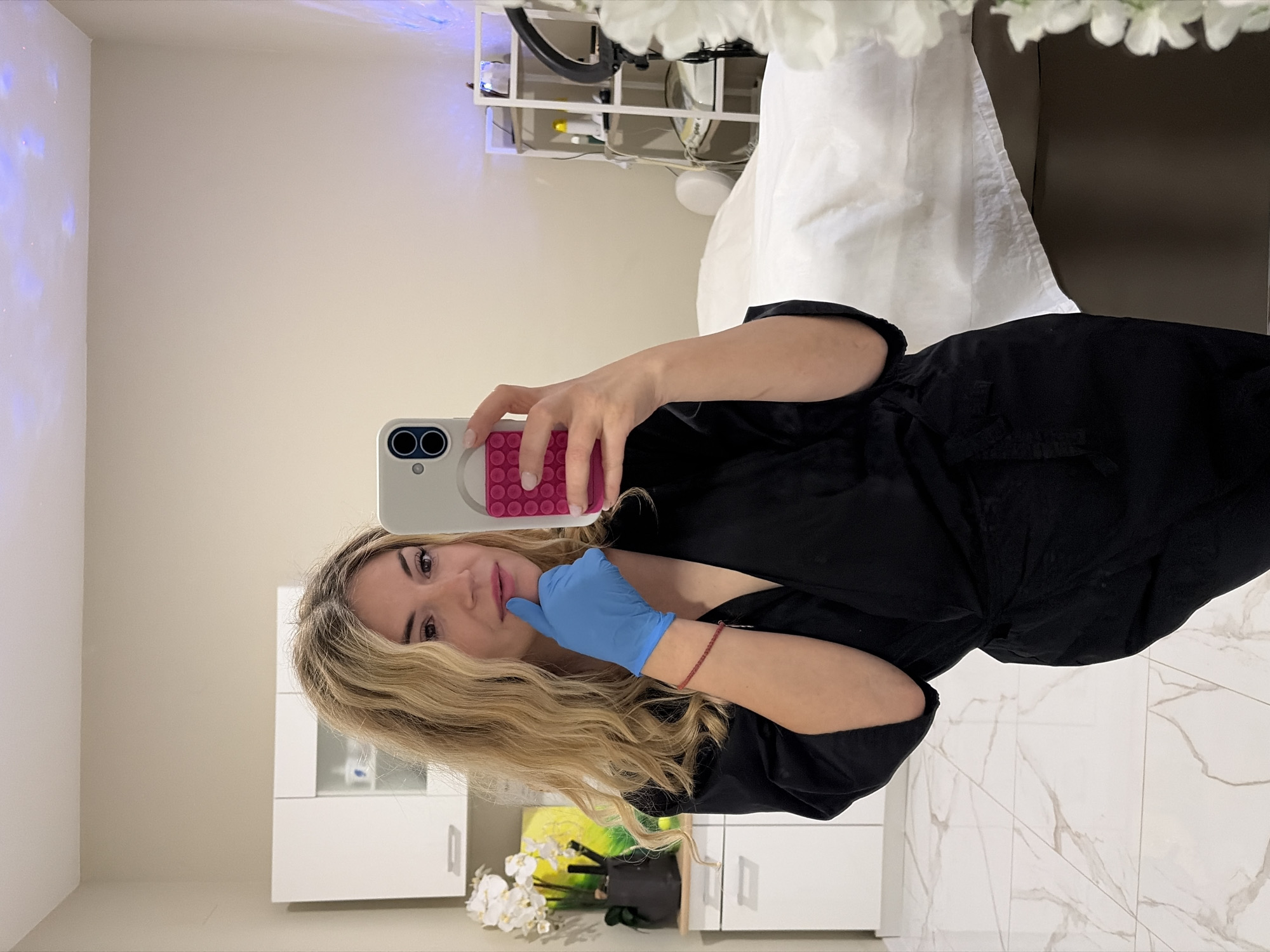A blonde woman in a black robe and blue surgical gloves poses for a selfie in what appears to be a medical or beauty clinic. She holds a phone with a pink and white case. The background includes white cabinets, a white medical bed, and a stand with what looks like medical supplies. The scene is well-lit with a mix of natural and artificial light. The floor is white with grey veining. It's impossible to determine the specific location in Budva, Montenegro, based solely on the image.