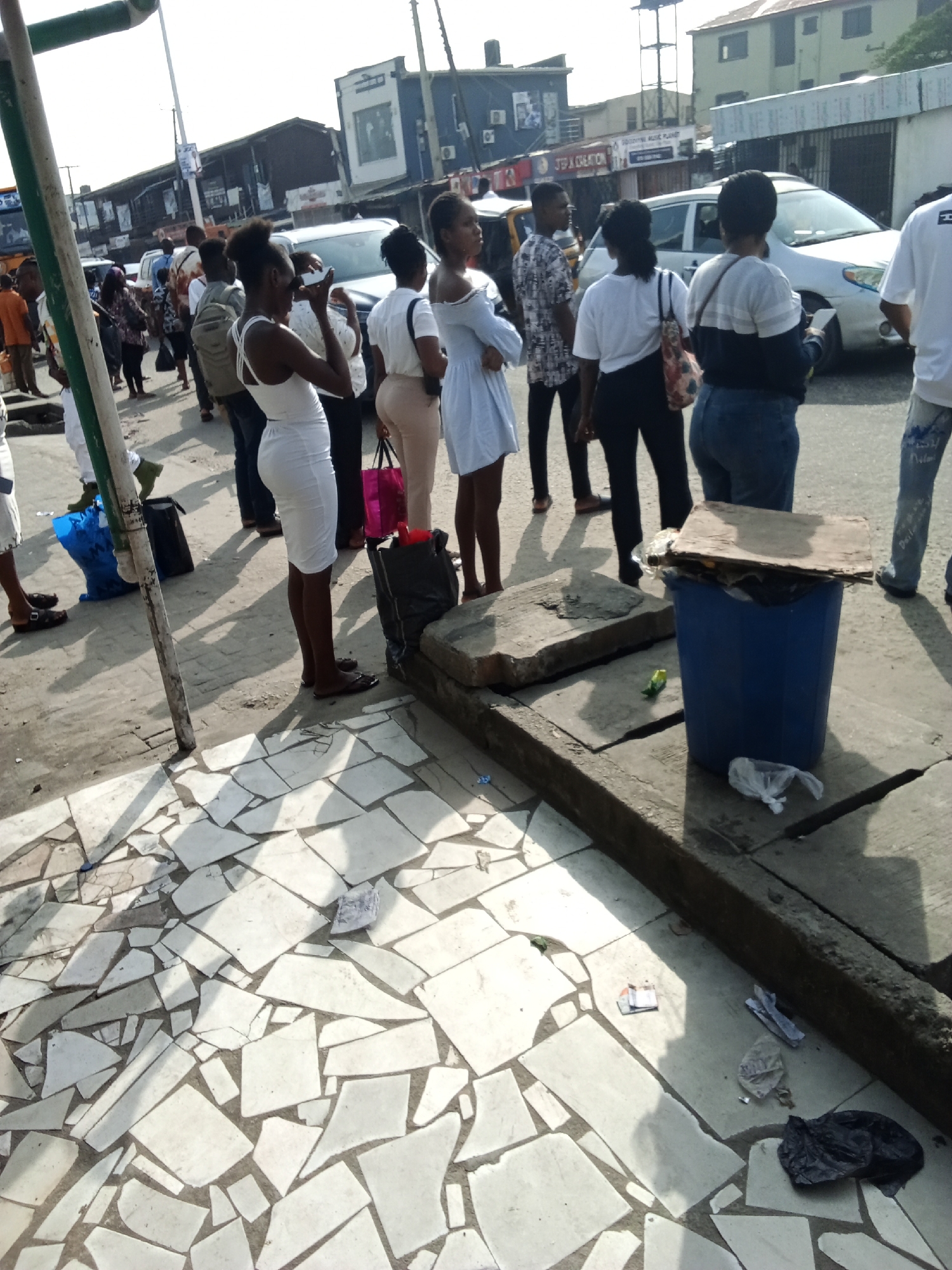 The image shows a street scene in Ado, Nigeria. A group of approximately 15 people are standing on a sidewalk. The sidewalk is made of broken white tiles and has litter scattered on it. A blue plastic trash can with a wooden plank on top sits near the edge of the sidewalk. Cars are visible in the background on a road. Buildings and businesses are also visible across the street.