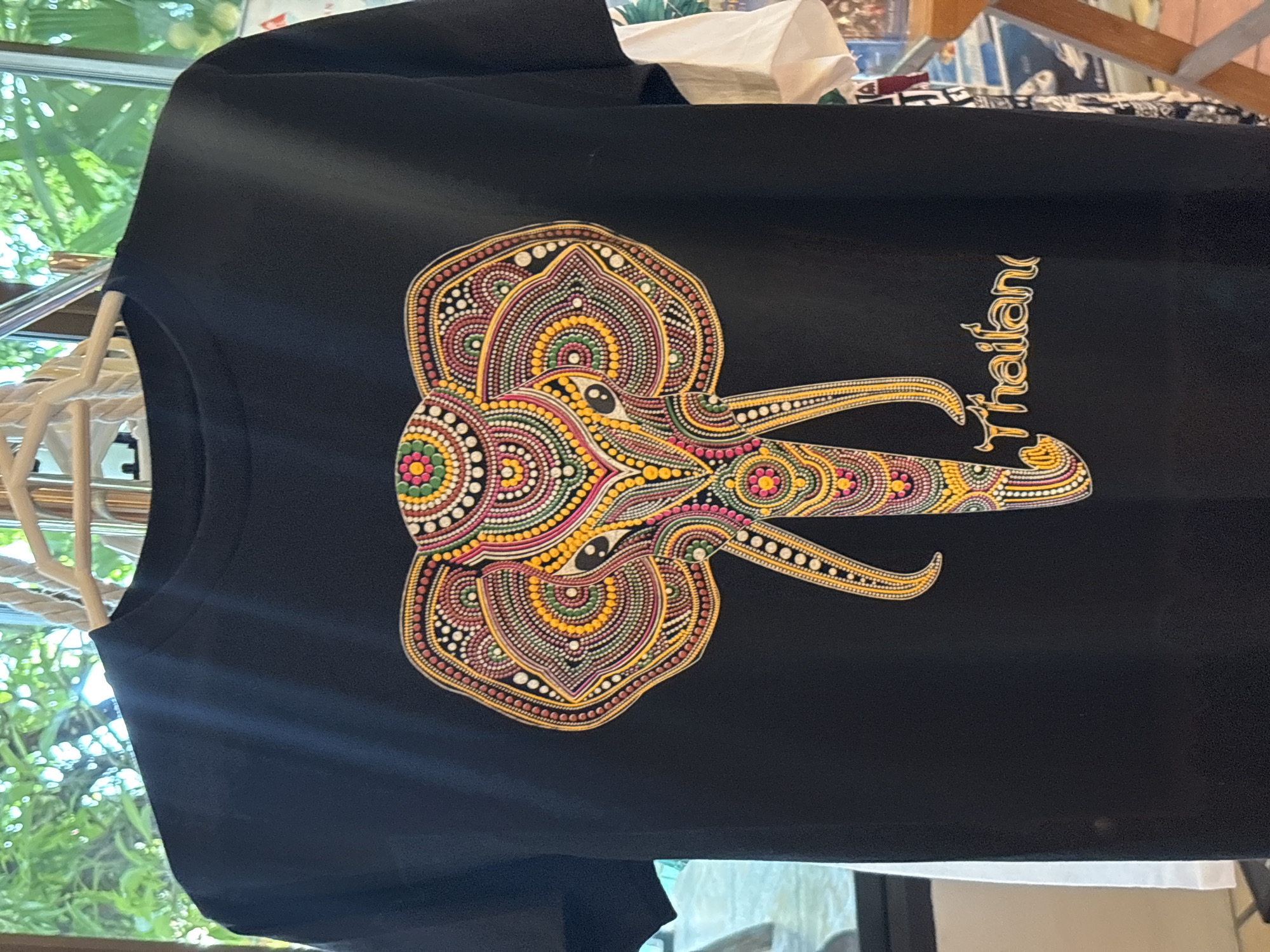 A black t-shirt with a colorful, dotted design of an elephant's head and the word "Thailand" written in yellow script is displayed. The t-shirt is hanging on a clothes hanger. In the background, out-of-focus greenery is visible through a window, suggesting an outdoor or semi-outdoor setting, possibly a shop or market stall.