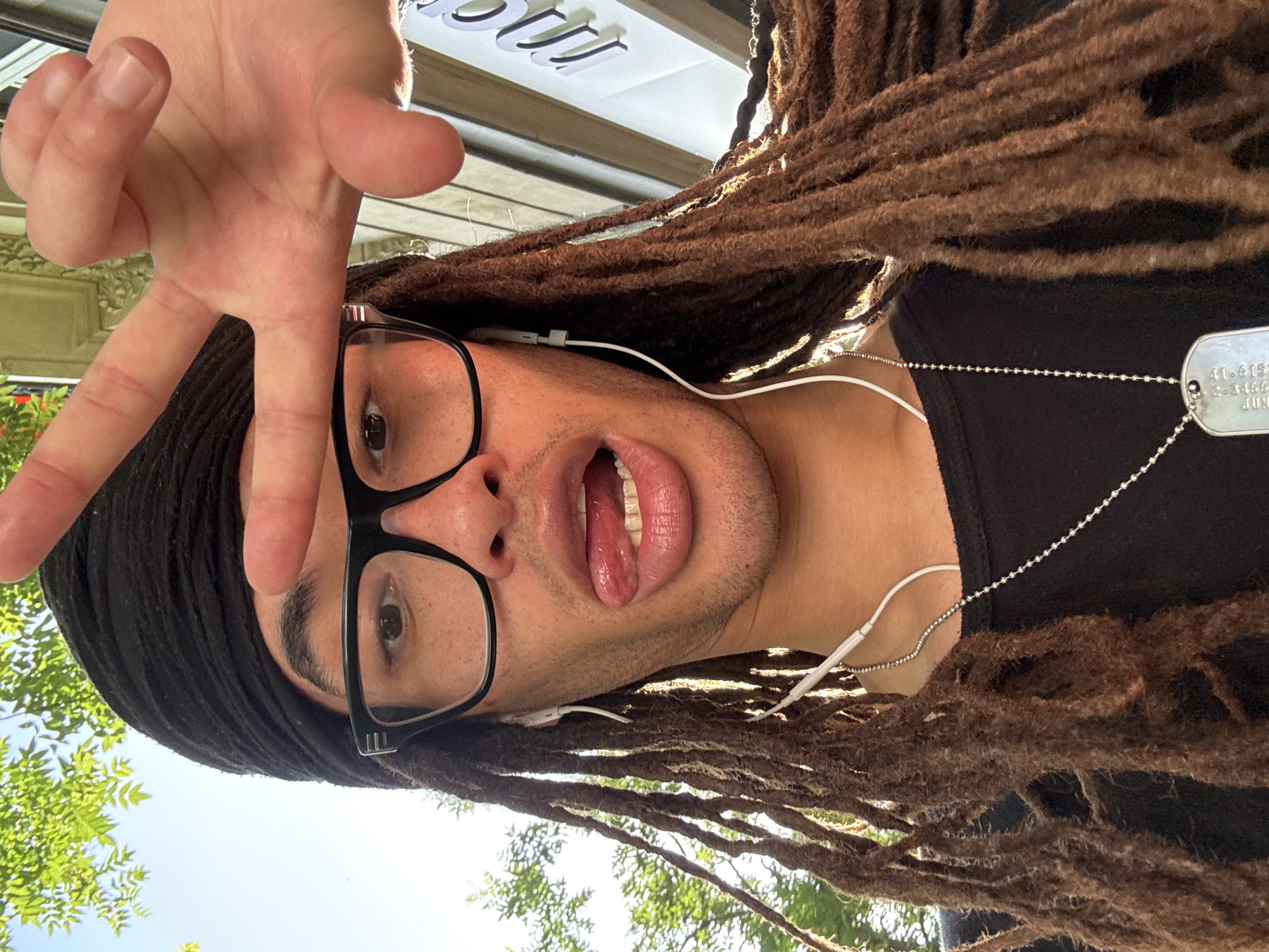 A male with brown dreadlocks, wearing a black shirt, black head wrap, and large-framed glasses, is seen in a low-angle selfie. He is displaying a hand gesture and sticking out his tongue. He is wearing earbuds and a dog tag necklace. The background indicates the location to be in Paris, France. There is a storefront sign with white lettering above him, and green trees are visible in the lower part of the image.