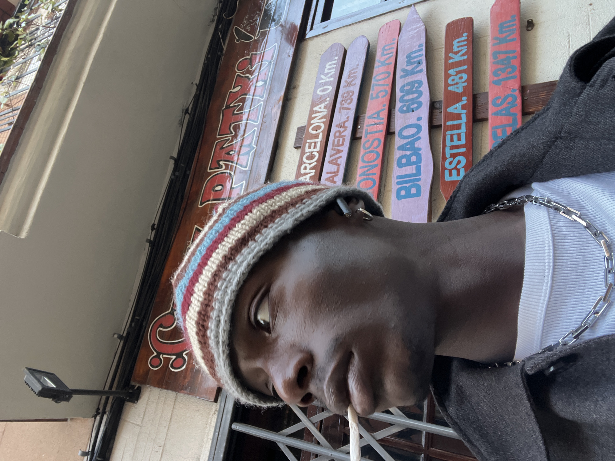 The image shows a man wearing a striped beanie, silver chain, and dark jacket, standing in front of a building displaying signs pointing to different cities and their distances in kilometers. The signs read "ARCELONA. 0 Km," "ALAVERA. 739 Km," "ONOSTIA. 570 Km," "BILBAO. 609 Km," "ESTELLA. 481 Km," and "FLAS. 1347 Km." The man is holding something thin, perhaps a cigarette, near his lips. The location is Barcelona, Spain, as indicated by the "ARCELONA. 0 Km" sign.