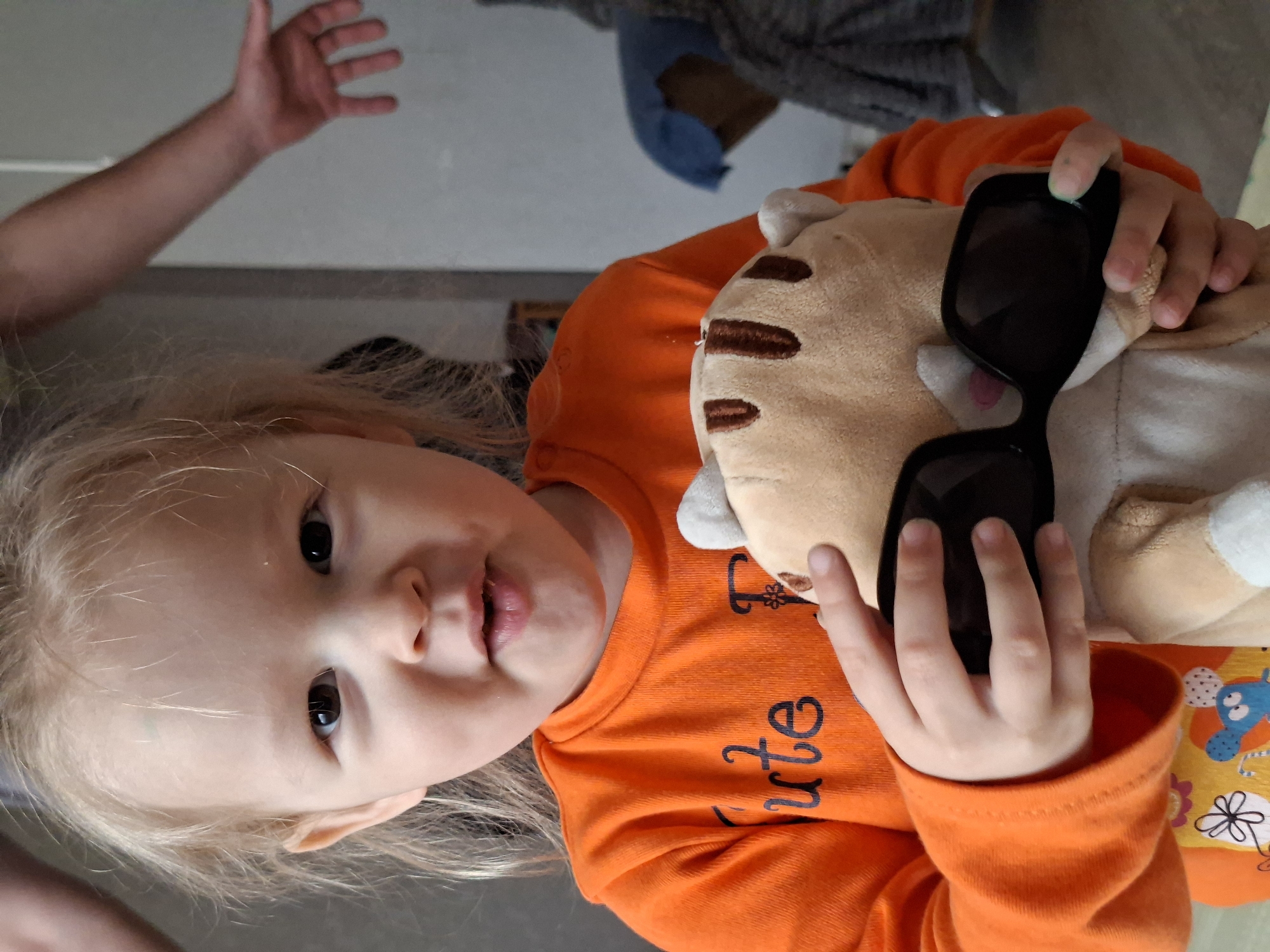 A candid photograph, rotated 90 degrees clockwise, features a young child, likely a toddler, with fair skin and light blonde hair. The child has dark, wide-open eyes and is looking directly at the camera with a slightly open mouth, giving a curious or playful expression.
The child is wearing a bright orange long-sleeved shirt. Visible in black stylized lettering on the chest of the shirt is the word "Cute". They are holding a light beige plush toy, which appears to be an animal like a cat or bear, with small brown embroidered stripes and ears. This plush toy is wearing a pair of black, oversized sunglasses, which the child's small hands are grasping along with the toy.
In the blurred background to the upper left, an adult's forearm and open hand are visible. The setting appears to be indoors, with generic, light-colored walls or surfaces and possibly a glimpse of another person's clothing. The overall impression is one of a playful and intimate moment. Below the child's orange shirt, a glimpse of another garment with a colorful pattern can be seen.