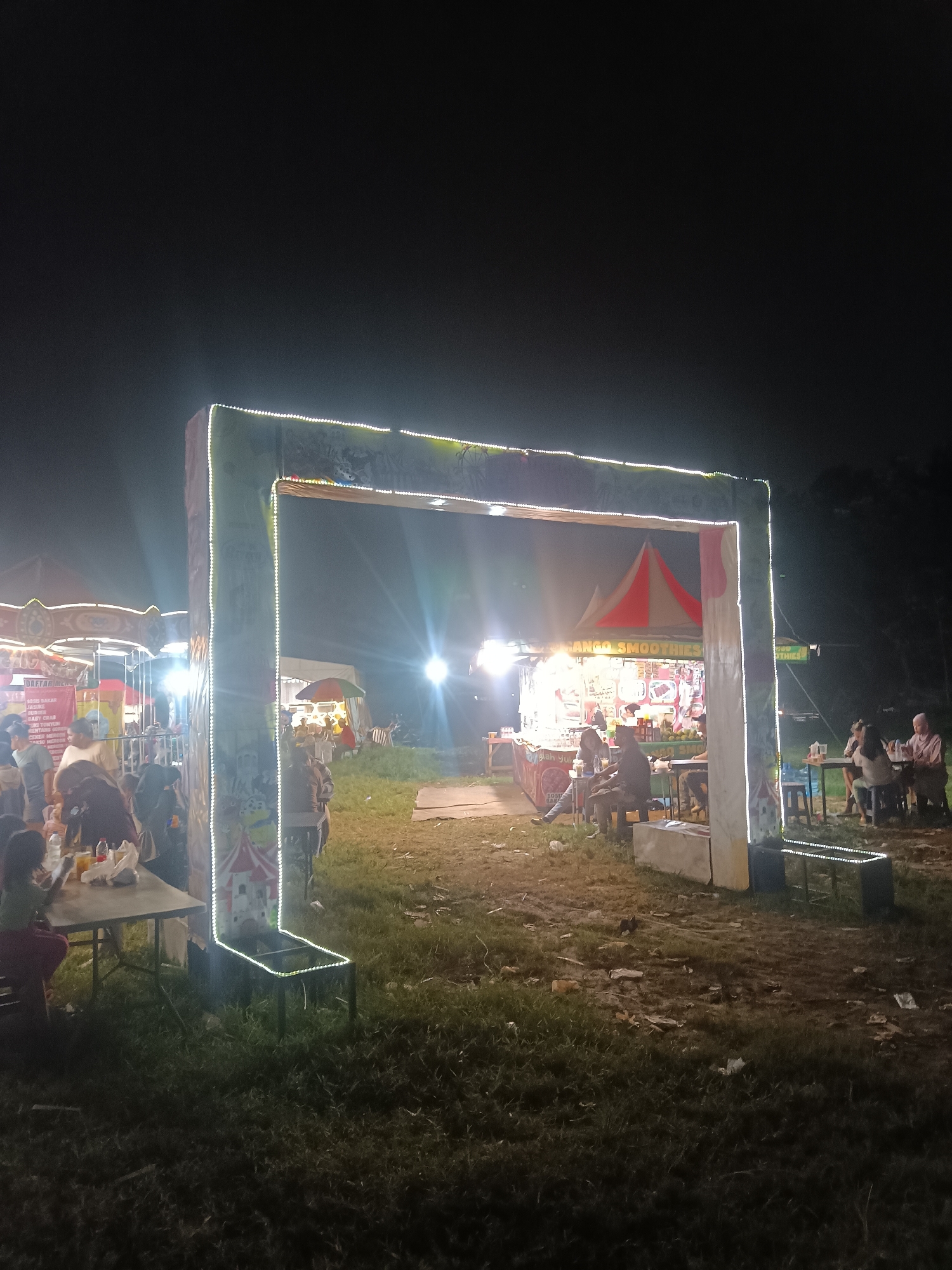 The image depicts a lively night market scene, likely in Depok, Indonesia. The main subject is a large, illuminated archway decorated with white fairy lights, serving as an entrance or focal point. Beyond the archway, a bustling food stall with a red and white striped awning is visible, advertising "MANGO SMOOTHIES". Several people are seated at tables in front of this stall and another in the background, enjoying food and drinks.
To the left of the archway, a carousel or carnival ride is partially visible, suggesting a festive atmosphere. People are scattered throughout the scene, some walking, others seated, engaged in social interaction. The ground appears to be a mix of grass and dirt, with some scattered debris, indicating an outdoor event. The time of day is clearly night, with artificial lights illuminating the scene and the sky dark. The overall mood is one of casual enjoyment and community gathering.
Visible text includes "MANGO SMOOTHIES" on the awning of the food stall. Another sign on the left appears to list food items like "SOTO AYAM", "BAKSO", "JAGUNG BAKAR", "BABY CHICKEN", "KENTANG GORENG", and "AYAM BAKAR". The lighting creates a vibrant and slightly blurry effect, enhancing the feeling of a lively night event.