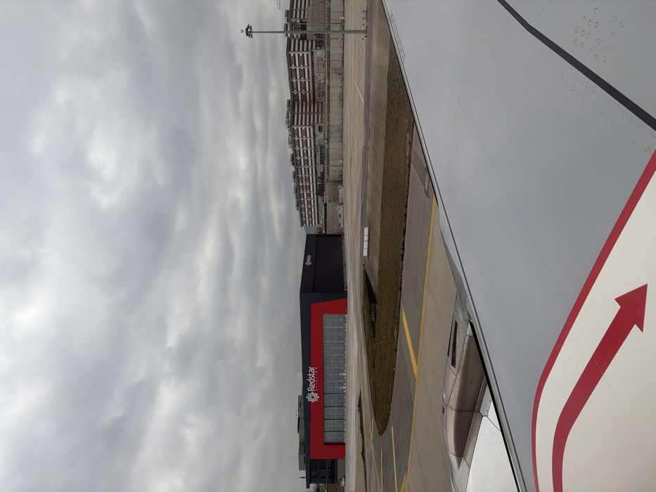 The image displays an exterior view from the right wing of an aircraft, positioned on an airfield apron at Sabiha Gökçen International Airport, located near Tuzla, Turkey. The aircraft's white wing, featuring a red arrow decal pointing aft-outboard and a section of its engine nacelle, occupies the lower right quadrant. Directly ahead, a large, modern hangar building belonging to "Redstar Aviation" is visible, characterized by a black structure with a prominent central red facade displaying the company's white logo and name. This building has multiple large, closed hangar doors. The foreground and midground consist of a grey asphalt taxiway or apron, marked with yellow lines, and interspersed with patches of dry, brown grass. In the background on the right, a multi-story industrial or commercial building complex, light brown in color with numerous horizontal bands and windows, is present. A tall, grey light pole stands in front of this complex. The sky is uniformly overcast with dense grey clouds. No individuals are visible, and the scene appears static.