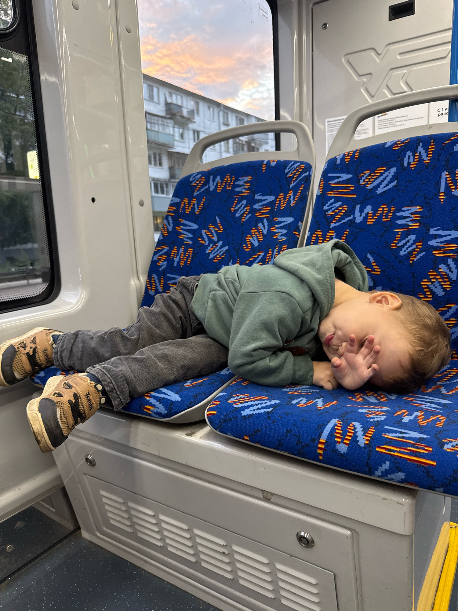 A young boy is sleeping on his side on a bus seat in Rusanovo, Russia. He is wearing a green hooded sweatshirt, gray pants, and brown and black sneakers. His face is turned away from the camera, and his hand is covering his eyes. The bus seats are blue with a pattern of orange and yellow zigzags. Outside the window, a building and a cloudy sky can be seen. The bus appears to be in motion, as suggested by the blur of the background. The lighting indicates it is either early morning or late evening.