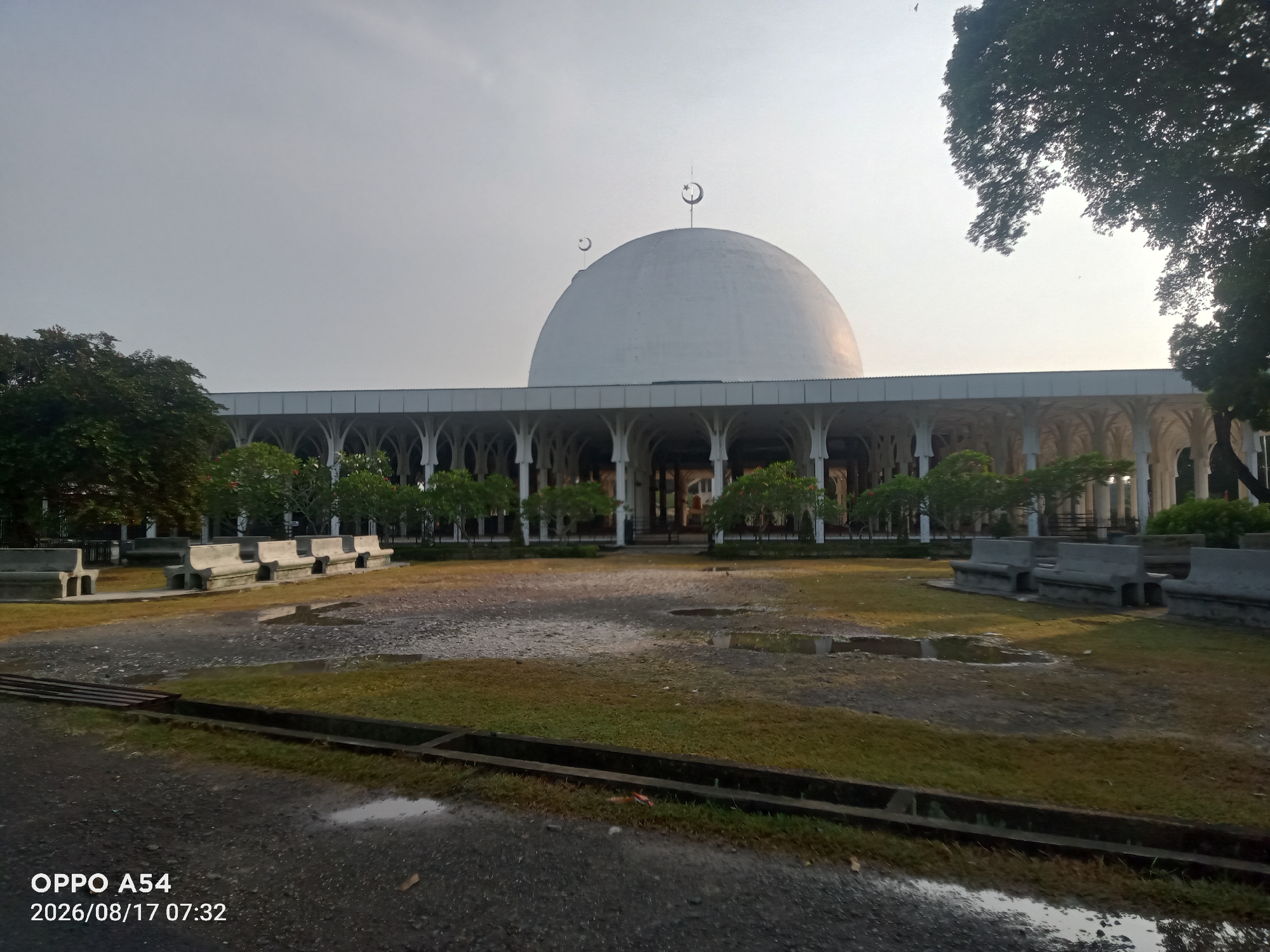 The image shows the exterior of the Great Mosque of Jambi, located in the city of Jambi, Indonesia. The mosque features a large white dome with crescent moons and stars adorning its peak. A colonnade of white arches surrounds the main building, with lush green trees and manicured lawns in the foreground. Several concrete benches are scattered throughout the courtyard, and puddles of water are visible on the paved ground, suggesting recent rainfall. The sky is overcast with a pale, hazy light, indicating it is likely early morning or late afternoon. The timestamp on the image indicates it was taken on August 17, 2026, at 07:32. There are no people visible in the image.