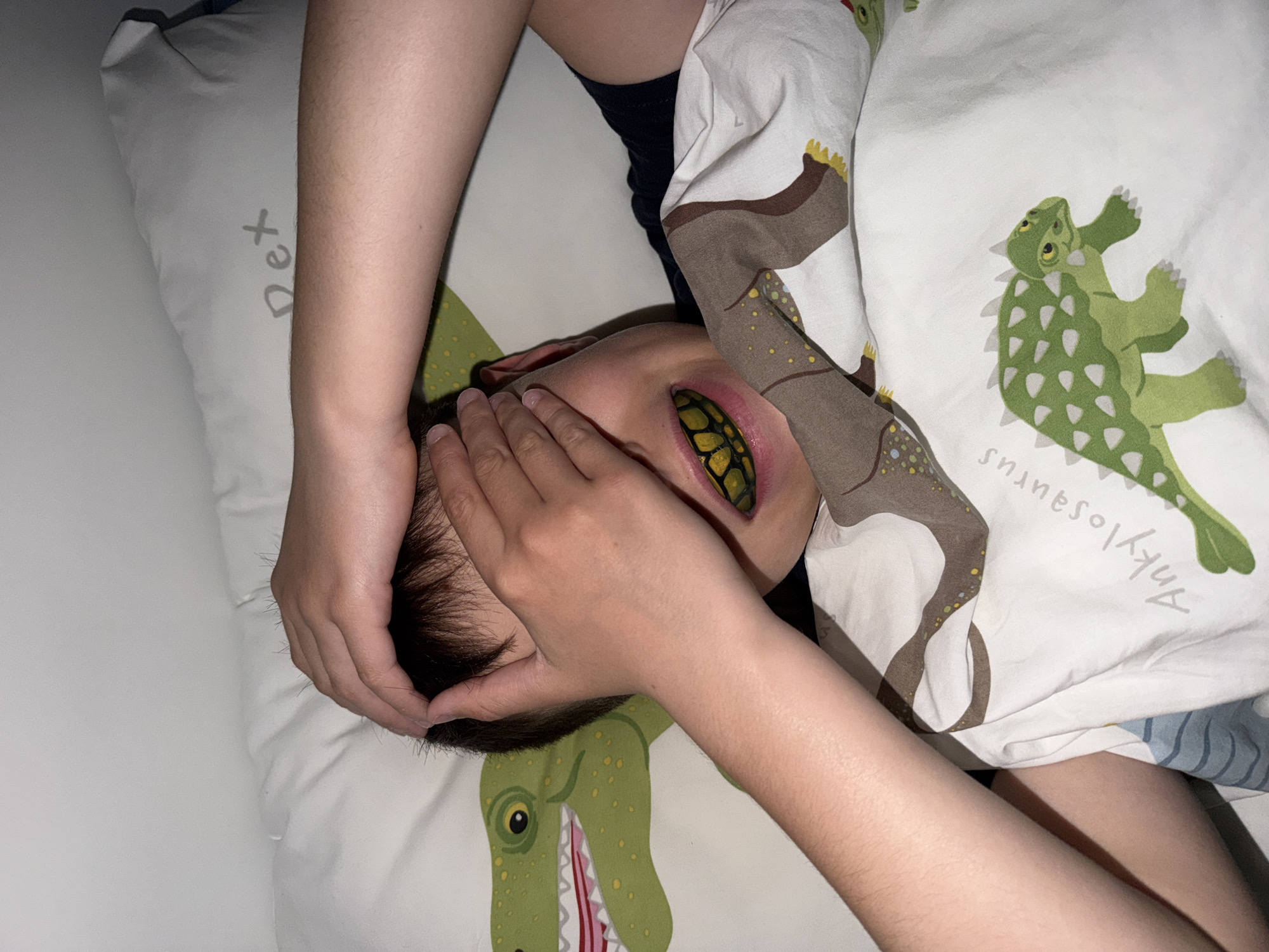 A person lies in bed, covering their eyes with their hands. They are partially covered by a dinosaur-themed bedsheet and pillow. The location is unspecified and cannot be determined to be in Votkinsk, Russia, or anywhere else.