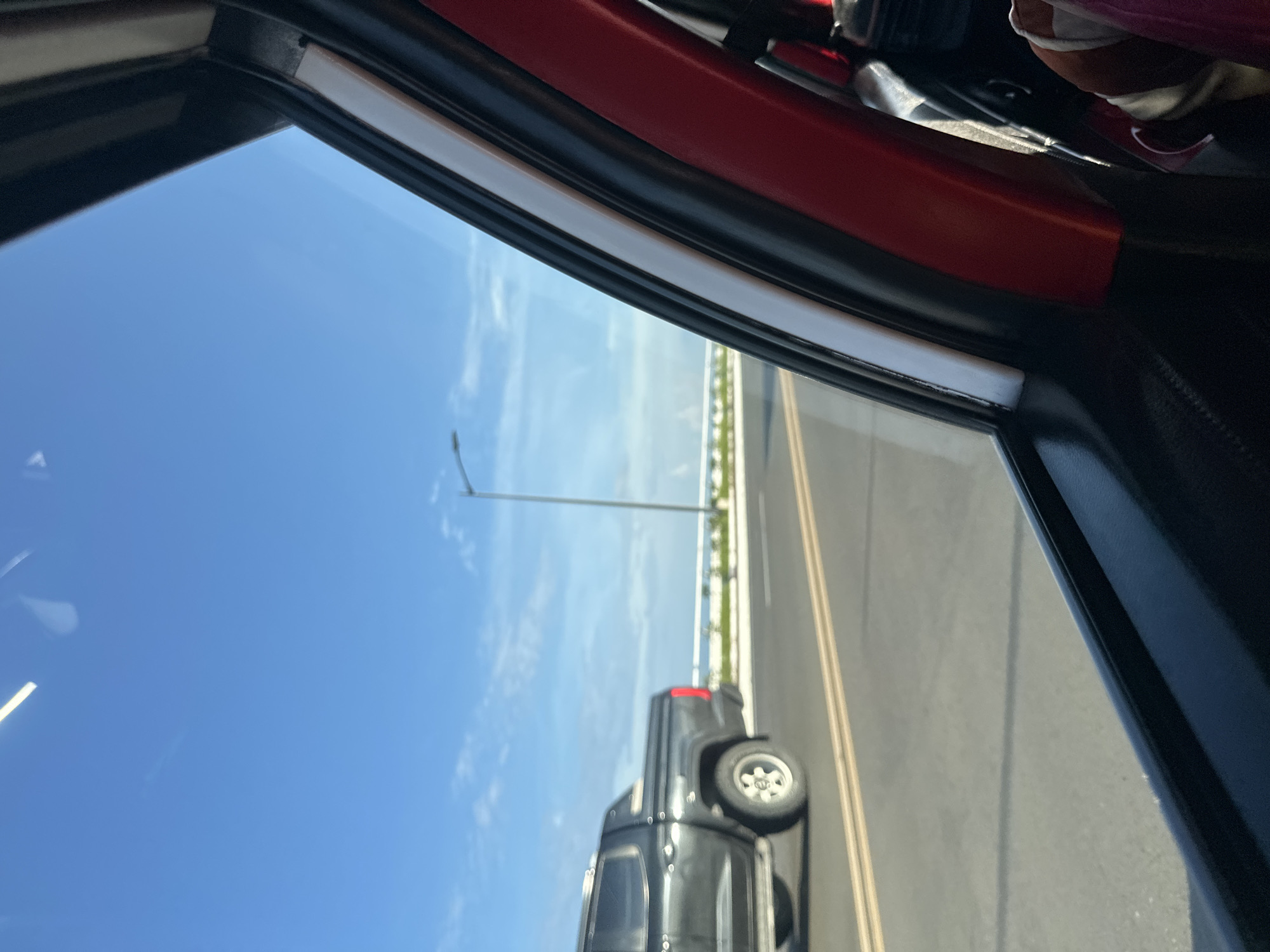 The image captures a roadside scene from the perspective of a moving vehicle on a clear day in Davao City, Philippines. The right side of the frame reveals the interior of the vehicle, specifically the dark red trim of the door pillar and a portion of the window frame. In the bottom right corner, a person's arm and shoulder, adorned with a striped shirt and pink fabric, are partially visible.
Outside the window, a multi-lane asphalt road is visible with two distinct solid yellow lines. A dark gray pickup truck, with its rear section and silver wheels in focus, is traveling on the road to the left of the viewer's vehicle. In the midground, a white concrete barrier or railing runs parallel to the road, suggesting a bridge or coastal highway. Beyond this barrier, a broad expanse of blue water, indicative of the ocean, meets the horizon. The sky above is a bright, clear blue with sparse, wispy white clouds. A solitary streetlamp pole with an extended arm stands in the distance on the right side of the road. Bright daylight illuminates the entire scene, casting subtle shadows on the road surface.