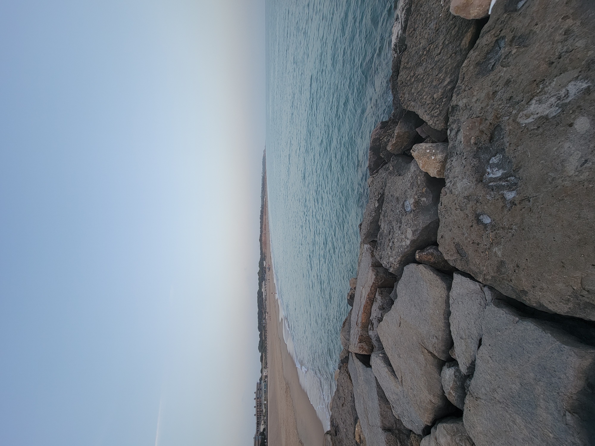 The image depicts a calm ocean with a sandy beach in the background, situated in Quarteira, Portugal. In the foreground, large, rough-hewn rocks form a breakwater, separating the viewer from the water. The sea is a serene turquoise, gently lapping the shore. The beach is wide and mostly empty, with a few scattered individuals visible near some buildings that line the coast. The sky is a soft, gradient blue, indicating it might be early morning or late evening. The overall atmosphere is peaceful and tranquil.