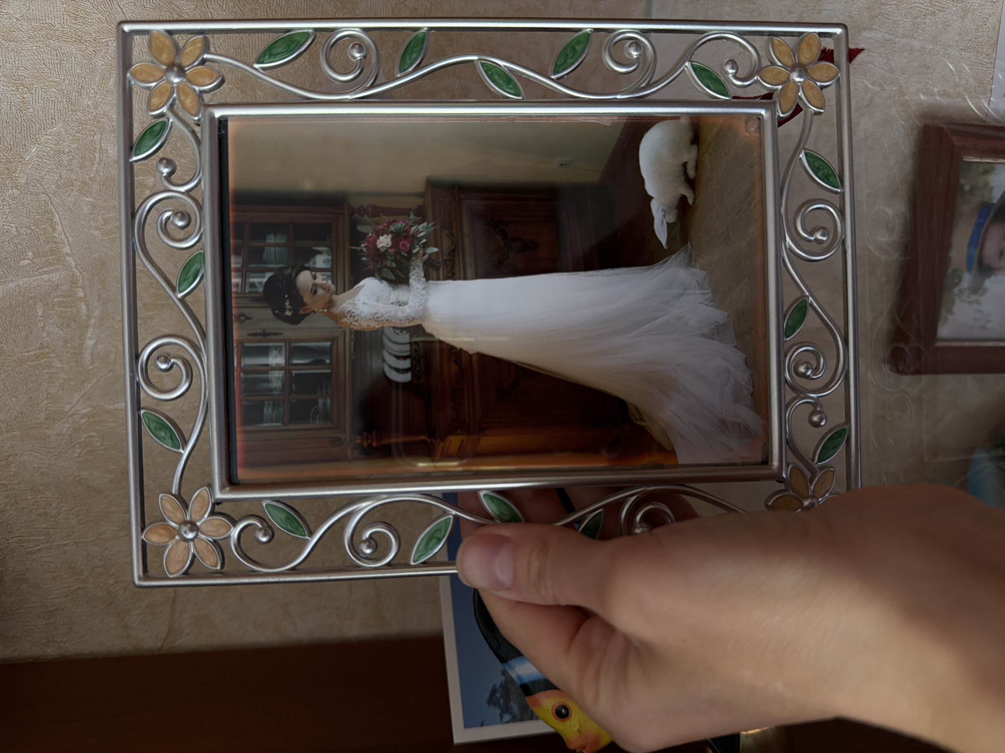 A person is holding a silver picture frame, which contains a photograph of a woman in a white wedding dress holding a bouquet. Behind the woman in the photo is dark wooden furniture and what appears to be a white cat. The frame is ornate with flower and leaf designs. Part of a hand is visible holding the frame. Behind the frame are a wall, another framed picture, and a card. The picture was not taken in Kamyshin, Russia.