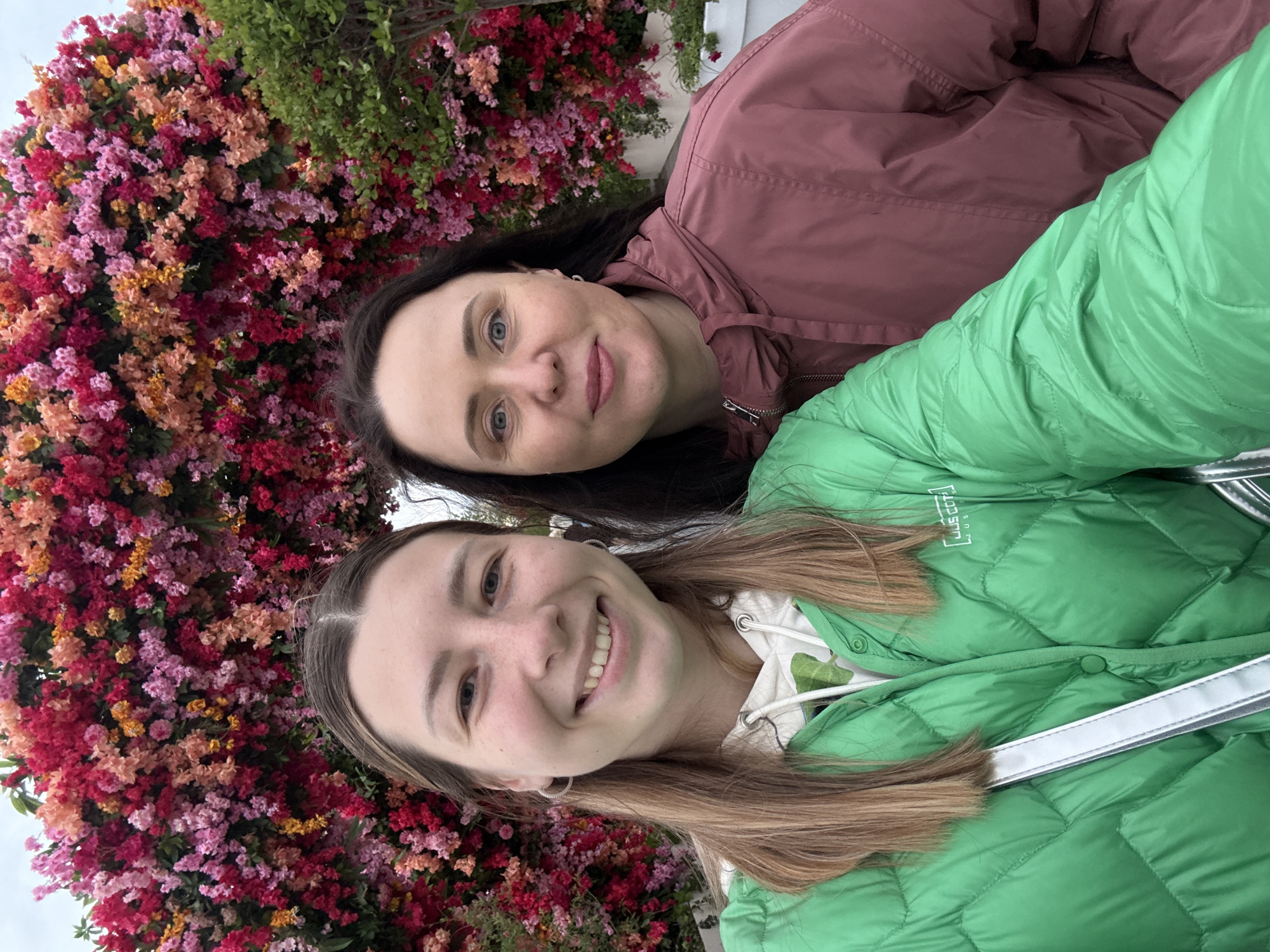 Two women pose for a selfie in front of a floral display. One woman has dark hair and is wearing a maroon jacket. The other has light brown hair and is wearing a green quilted jacket. The floral display behind them features a variety of colors including red, pink, orange, and yellow. Based on the types of flowers and background elements, it is likely the photo was taken in the city of Moscow, Russia.