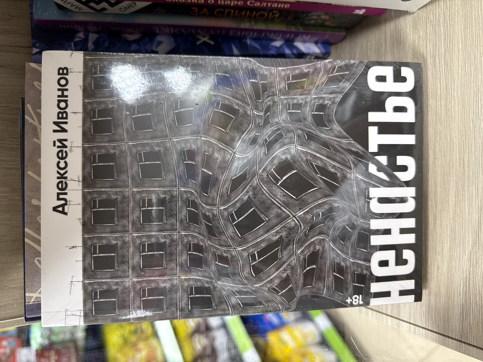 An overhead, slightly angled shot captures a shrink-wrapped book by Russian author Alexei Ivanov, titled "ненастье" (Nenastye, meaning "Bad Weather" or "Foul Weather"), resting on a light-colored wooden shelf or table. The book's cover features a striking, surreal image of a distorted, wavy grid resembling a building facade with dark windows, rendered in shades of grey and black, conveying an unsettling atmosphere. An "18+" age restriction is visible on the bottom right of the cover.
Behind and above this main book, several other books are stacked or arranged on shelves. Partially visible titles include "о царе Салтане" (About Tsar Saltan), "ЗА СПИНОЙ" (Behind the Back), "OMEP" (likely a partial title, possibly 'Homer'), and "МАЙ" (May). Some other faint and truncated text, such as "ПАРКНЫЙТИ МАЯКЕ" and "UPPOR," is also visible on the spines of these background books. The background below the shelf is blurred, showing colorful shapes that suggest other items or products in an indoor setting, likely a bookstore or a home library in Moscow, Russia. The scene is static, capturing the arrangement of books.