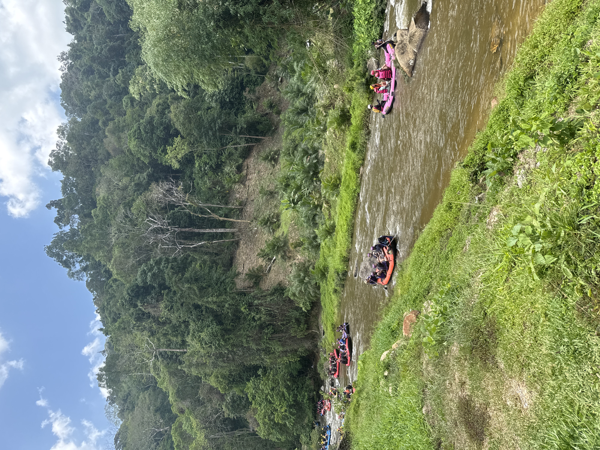 The image shows a river scene in Tambon Song Phraek, Thailand, filled with multiple rafts. A vibrant green forest covers the land on the left. The water is murky, and the rafts are spaced out along the river's length. Some rafts are pink, others orange or black, each carrying several individuals. People appear to be paddling. The landscape is lush with vegetation, and the sky shows a patch of blue.