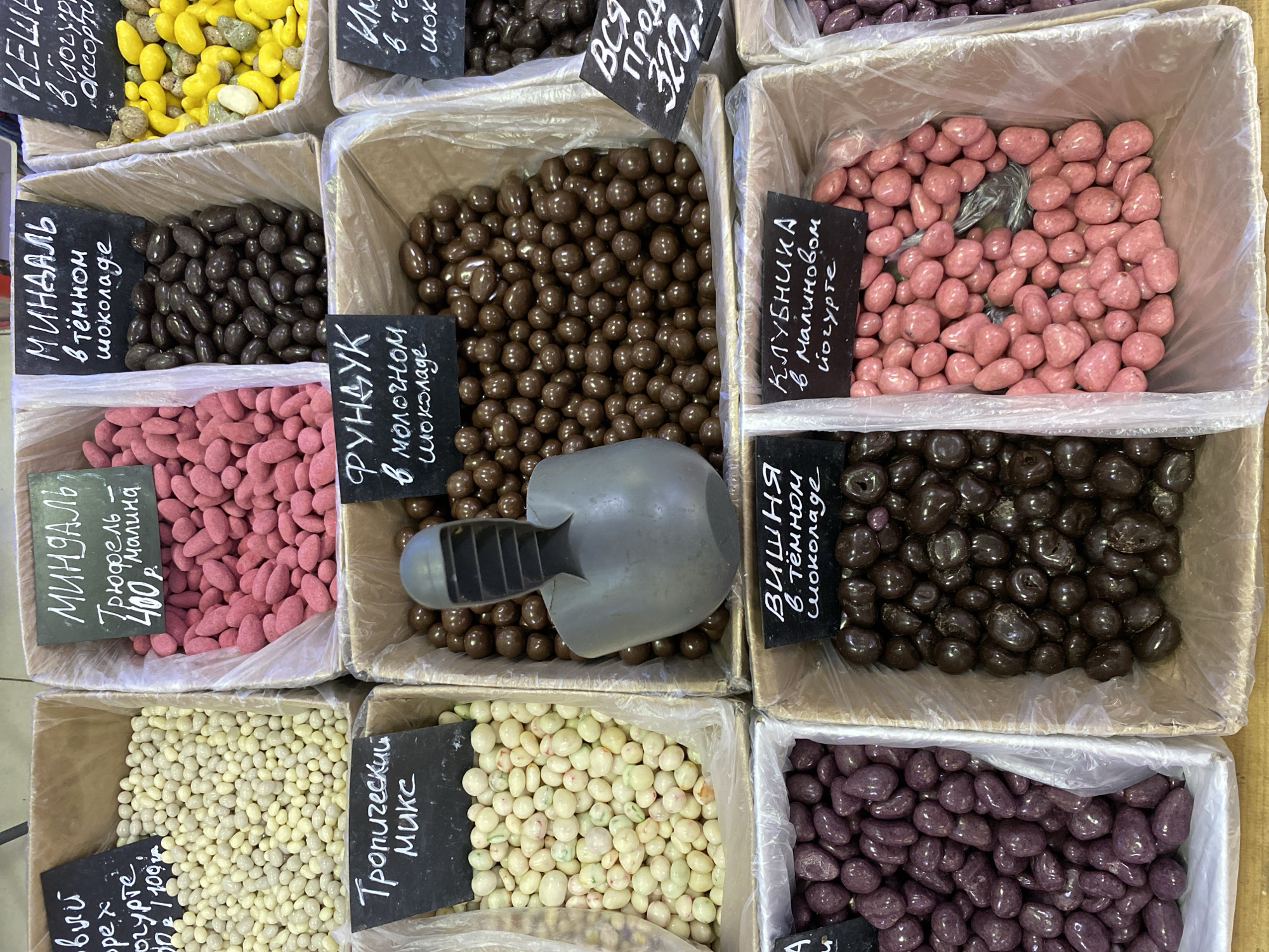 The image shows a display of various types of chocolate-covered nuts and other confections in a store, likely in the city of Chaikovsky, Russia, given the Russian language on the labels. The items are arranged in individual boxes or containers, some lined with plastic bags. There are nuts coated in different types of chocolate and yogurt. The colors of the coatings vary from yellow, white, pink, brown, and dark purple. Some of the labels indicate the type of nut and chocolate, such as almond in dark chocolate and hazelnut in milk chocolate, as well as the price per weight. A grey plastic scoop rests in one of the boxes filled with brown chocolate-coated nuts. The containers appear to be arranged in rows, suggesting a market or confectionery shop setting.