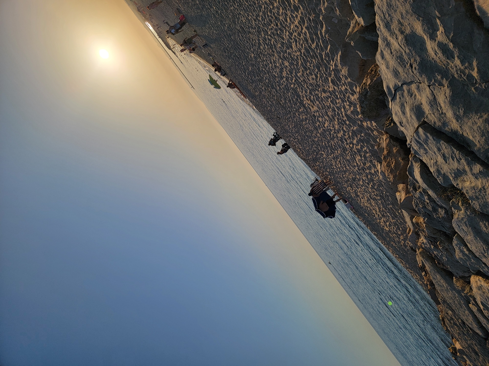 The image captures a beach scene in Quarteira, Portugal, during sunset. The sun is low in the sky, casting a warm, golden glow over the sand and sea. The sky transitions from a clear blue at the top to a pale yellow near the horizon.
The foreground is dominated by a rocky embankment leading down to the sandy beach. The sand itself shows the texture of small ripples, indicating the gentle lapping of waves.
Along the beach, several people are visible. Some are scattered further up the shore, near what appears to be a walkway or promenade. Closer to the water's edge, a few individuals are seated or lying on the sand, some under a blue umbrella. A few other beachgoers are seen walking or standing. A person is also visible in the water.
The sea is calm and reflects the colors of the sky. The overall impression is one of a peaceful evening at the beach.