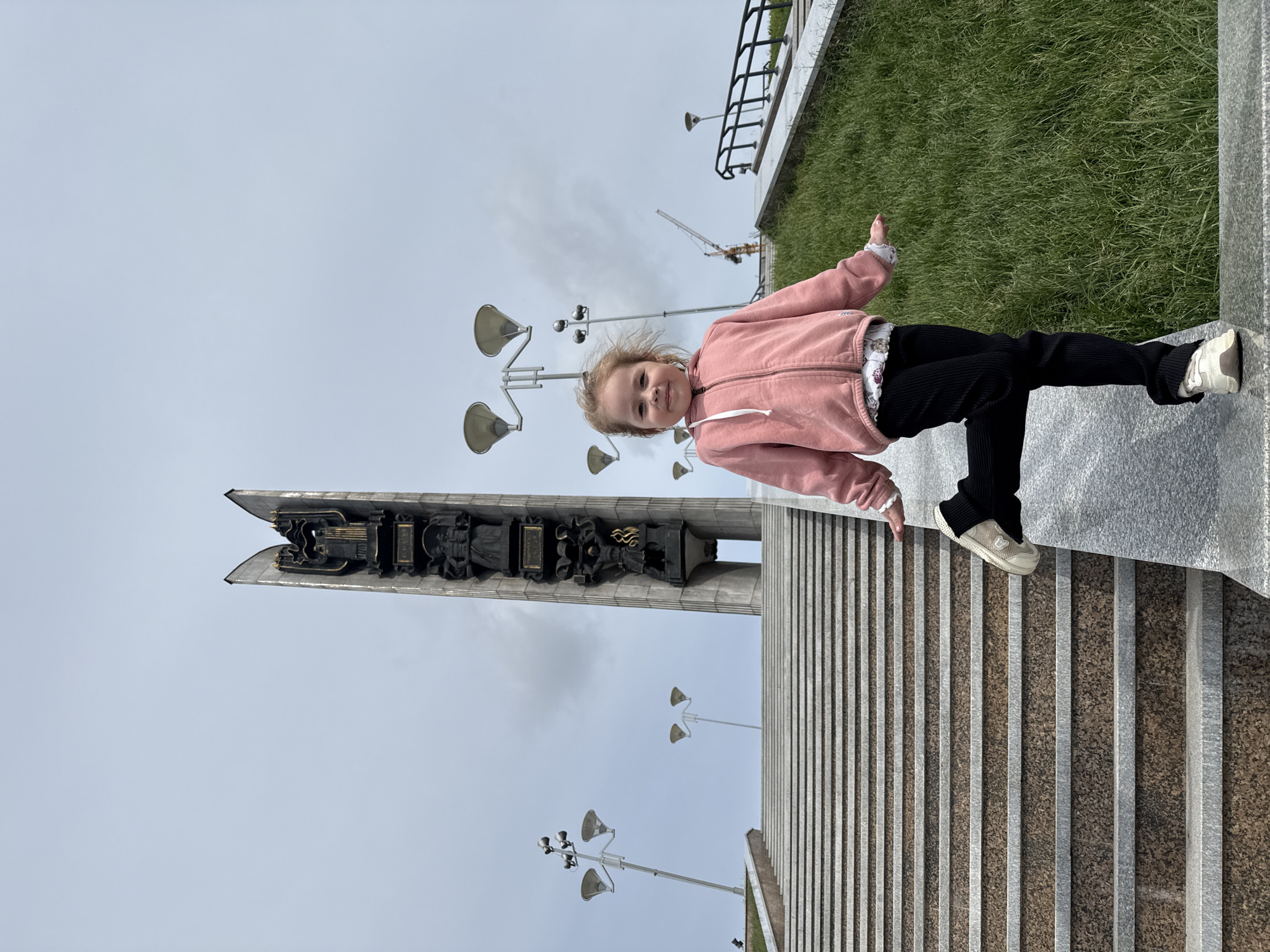 A young girl stands smiling brightly at the camera in a vibrant outdoor setting. She is positioned towards the right side of the frame, balancing with one foot on a wide granite stair and the other slightly raised, suggesting a playful pose. She wears a pink hooded jacket, black ribbed pants, and white sneakers with gray accents. Her light-colored hair is slightly windswept, hinting at a gentle breeze.
Behind the girl, to the left, stands a tall, striking monument or obelisk made of gray stone. It features intricate dark relief sculptures depicting historical or industrial themes, including figures and what appears to be a stylized weapon or industrial machinery. The monument's apex has a distinctive, sharp-edged design.
The surrounding area includes several multi-headed streetlights with broad, disk-shaped lamps. In the background, against an overcast sky, a construction crane is visible, indicating ongoing development. To the girl's right, a patch of green grass is bordered by a black metal railing and a concrete wall. The wide stairs are composed of alternating smooth gray and textured brownish-gray granite bands.
The scene takes place during the daytime under a cloudy sky, suggesting moderate weather. The location is consistent with a public space or memorial in Izhevsk, Russia. The girl's cheerful expression conveys a sense of happiness or enjoyment. No legible text is visible within the image.